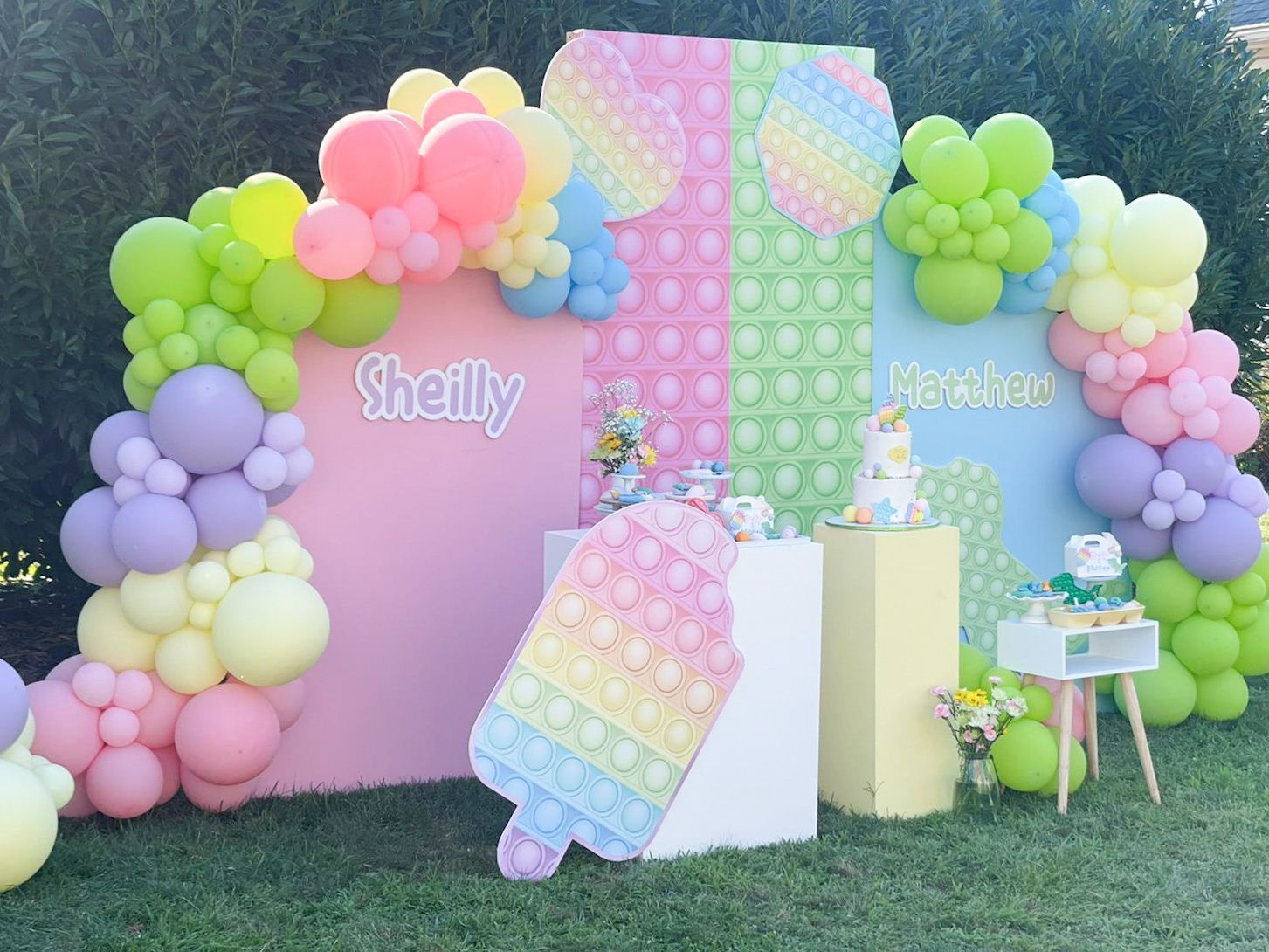 Party backdrop with Pop It theme: pastel colors, balloons, cake, and two names.