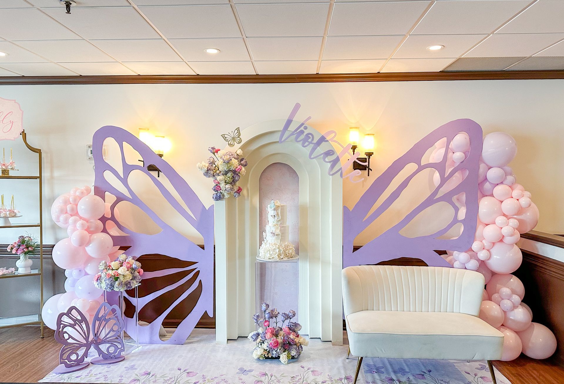 Butterfly-themed event backdrop with purple wings, balloons, floral arrangements, cake, and a white sofa.