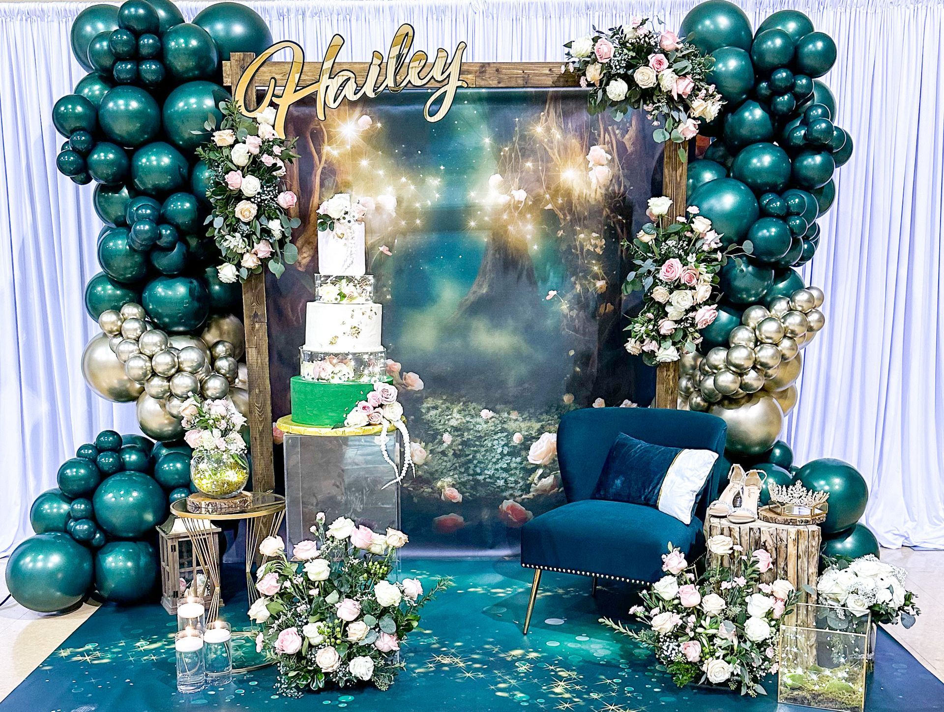 Emerald green party backdrop with balloons, cake, chair, and floral arrangements.
