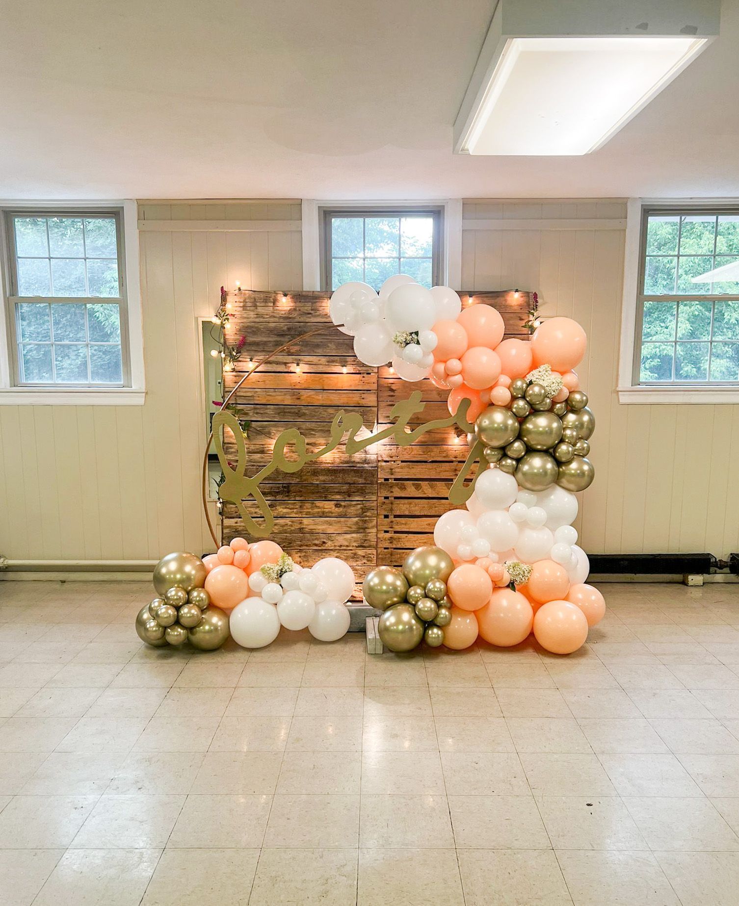 Balloon arch with gold, peach, and white balloons, in front of a wood backdrop with lights.