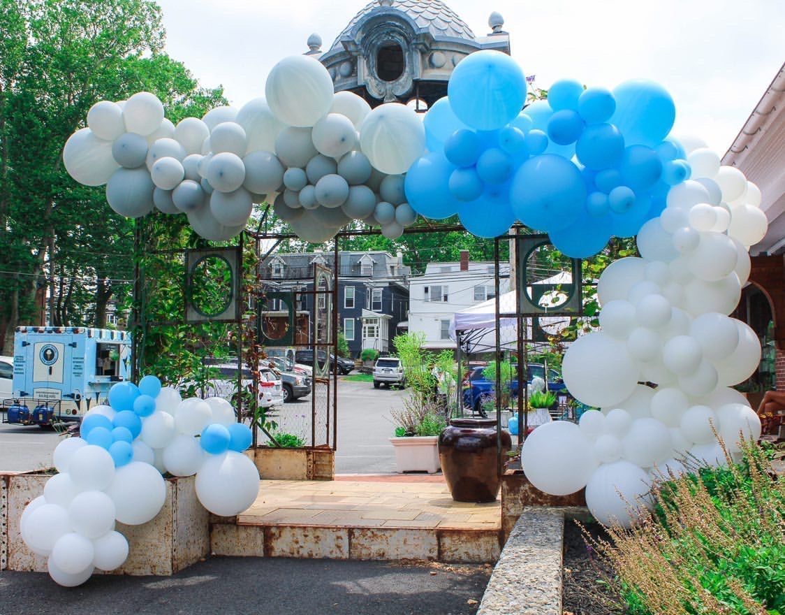 An archway decorated with blue, white, and gray balloons. In the background, a street with buildings.