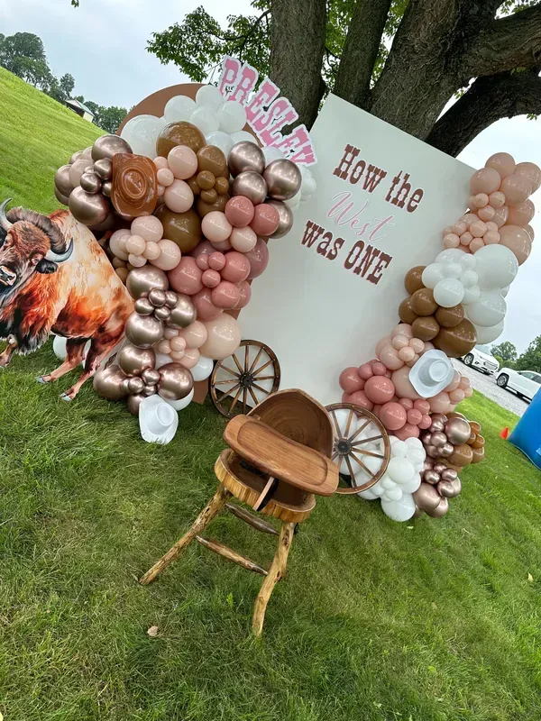 Cowgirl-themed first birthday party decorations: balloon arch, sign, high chair, and bison cutout on a grassy hill.