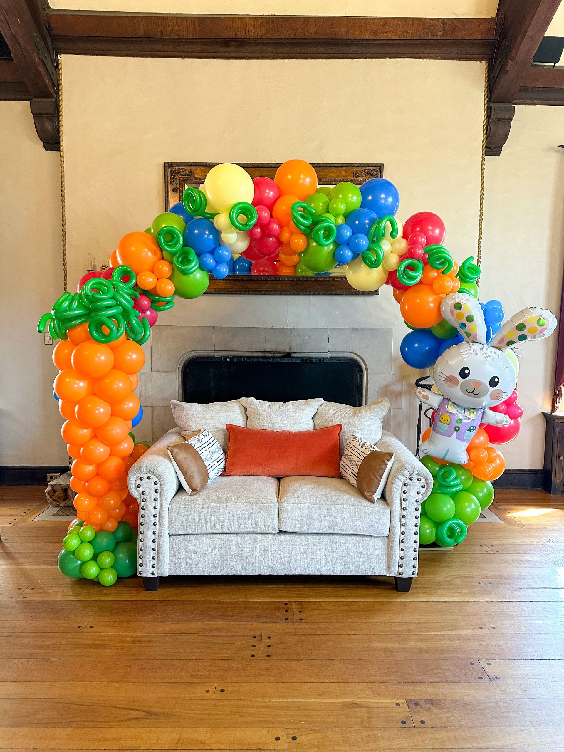 Easter balloon arch with carrot columns, bunny, and sofa in front of a fireplace.