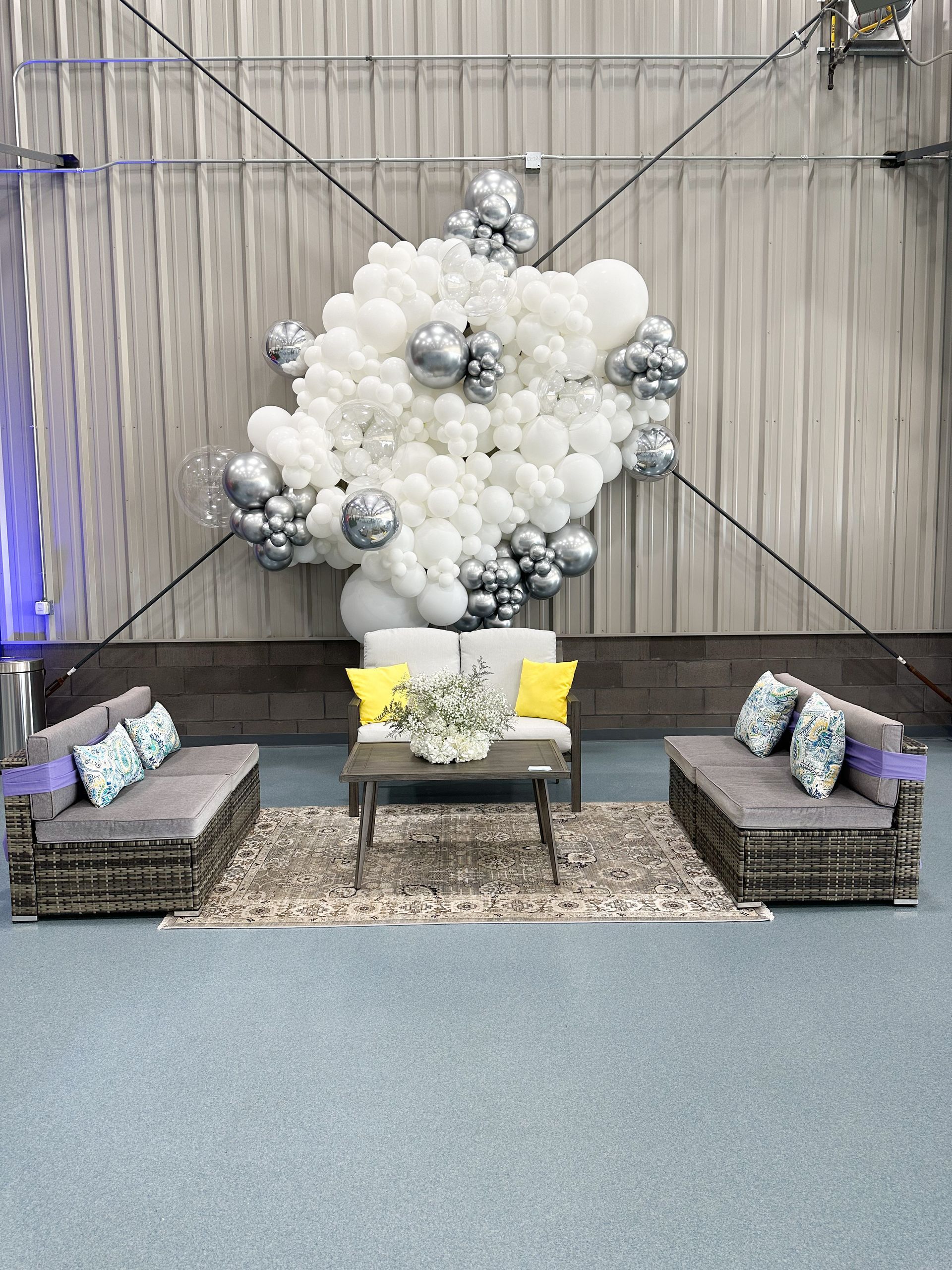 Lounge area with gray sofas, a rug, a coffee table, and a white and silver balloon arrangement against a gray wall.