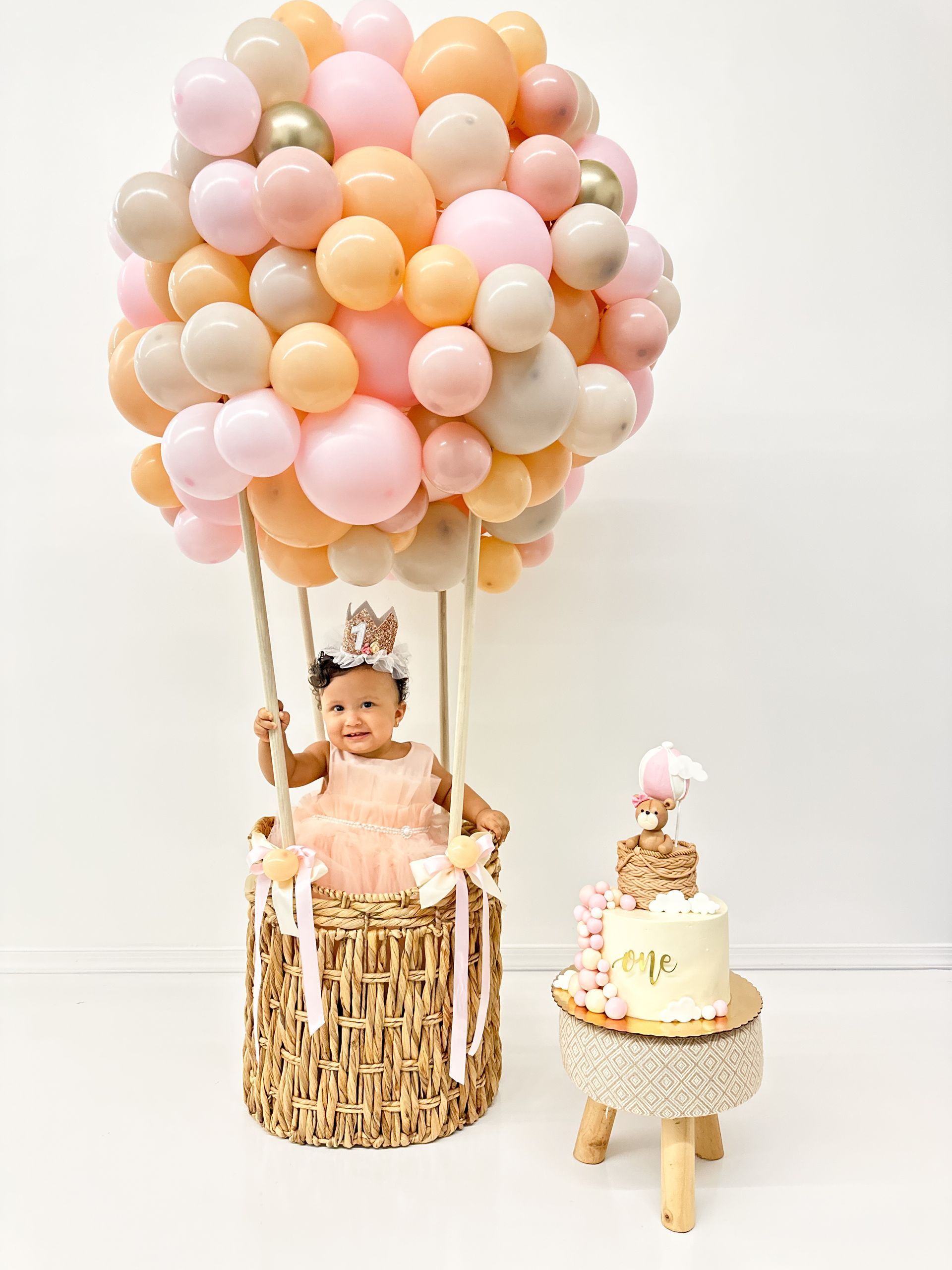 Girl in hot air balloon basket with pink and gold balloons, cake on small table.