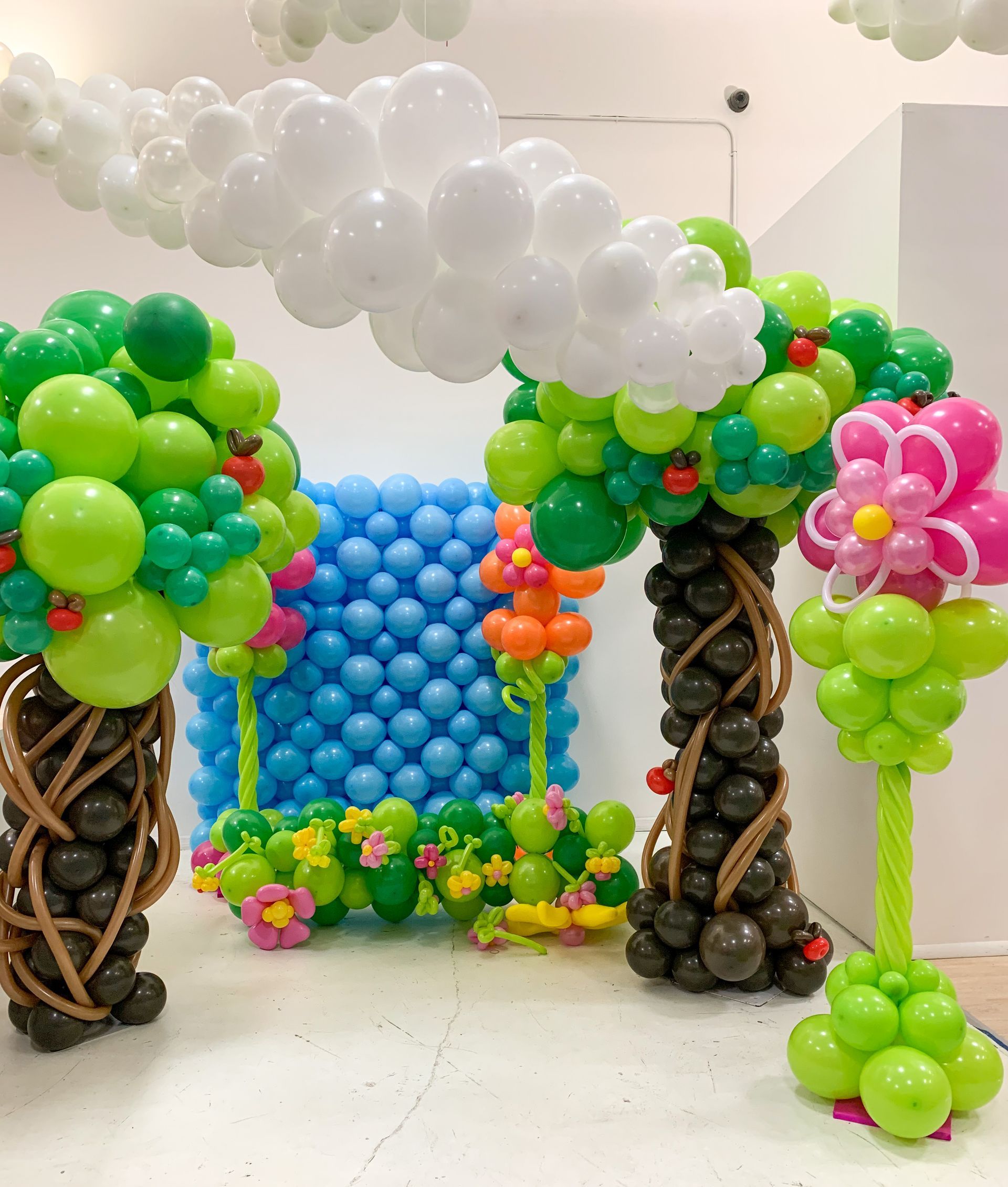Balloon art display: trees, flowers, and arch with white clouds; blue backdrop. Green, brown, pink, and white balloons.