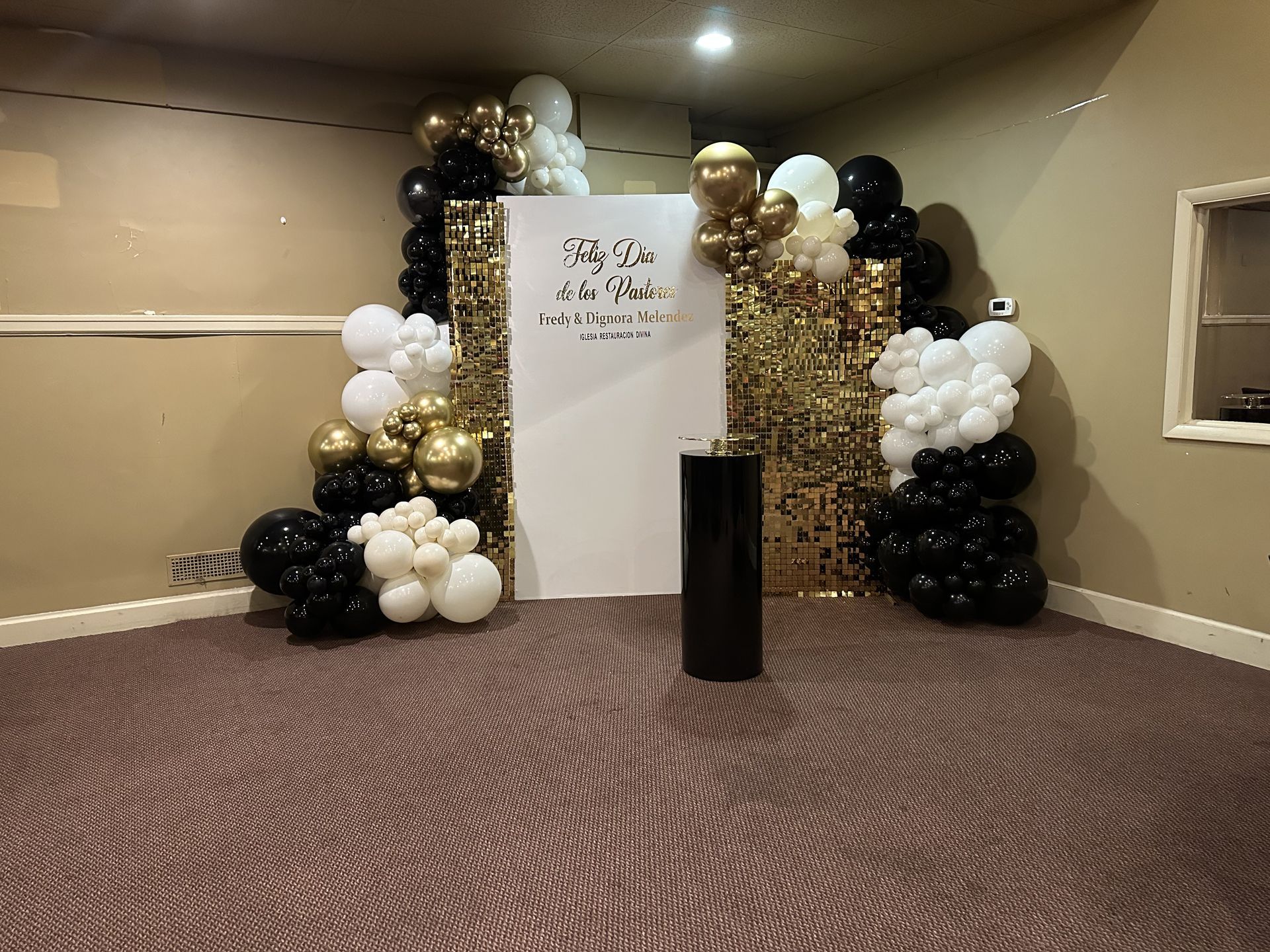 Black, white, and gold balloon arch decorates a room corner with a white sign, against a gold fringe backdrop.