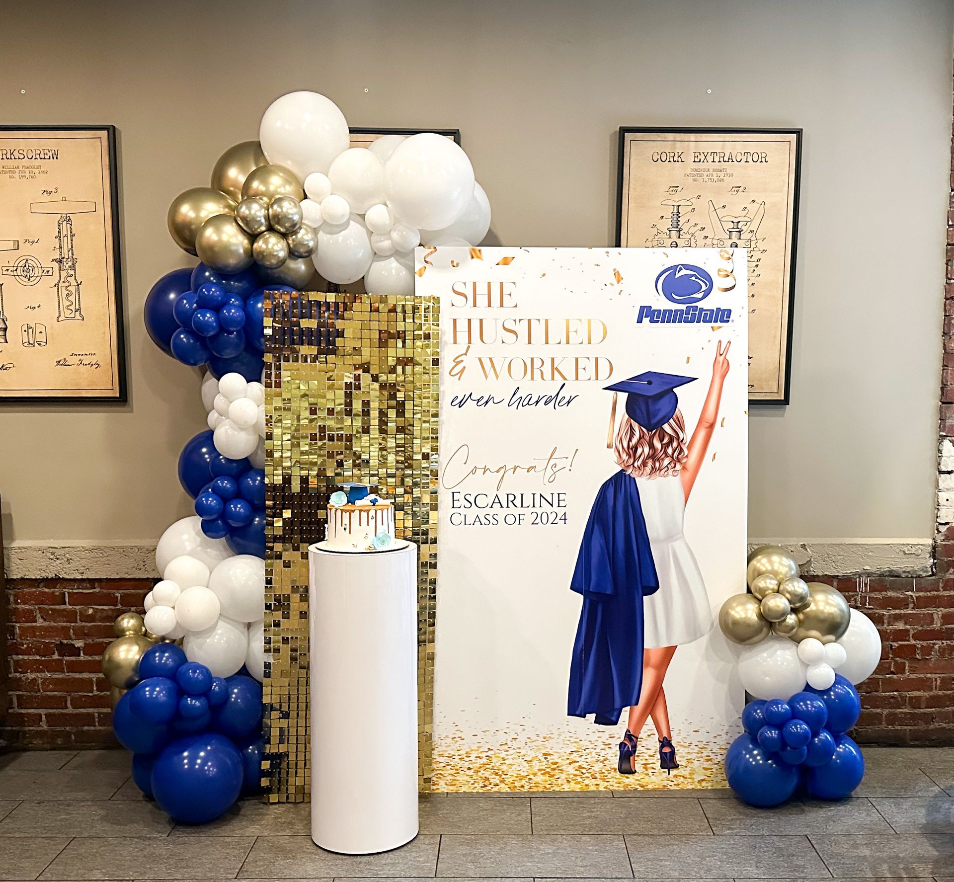 Graduation party display: balloons, sign with illustration of graduate, cake on a pedestal, and golden wall.
