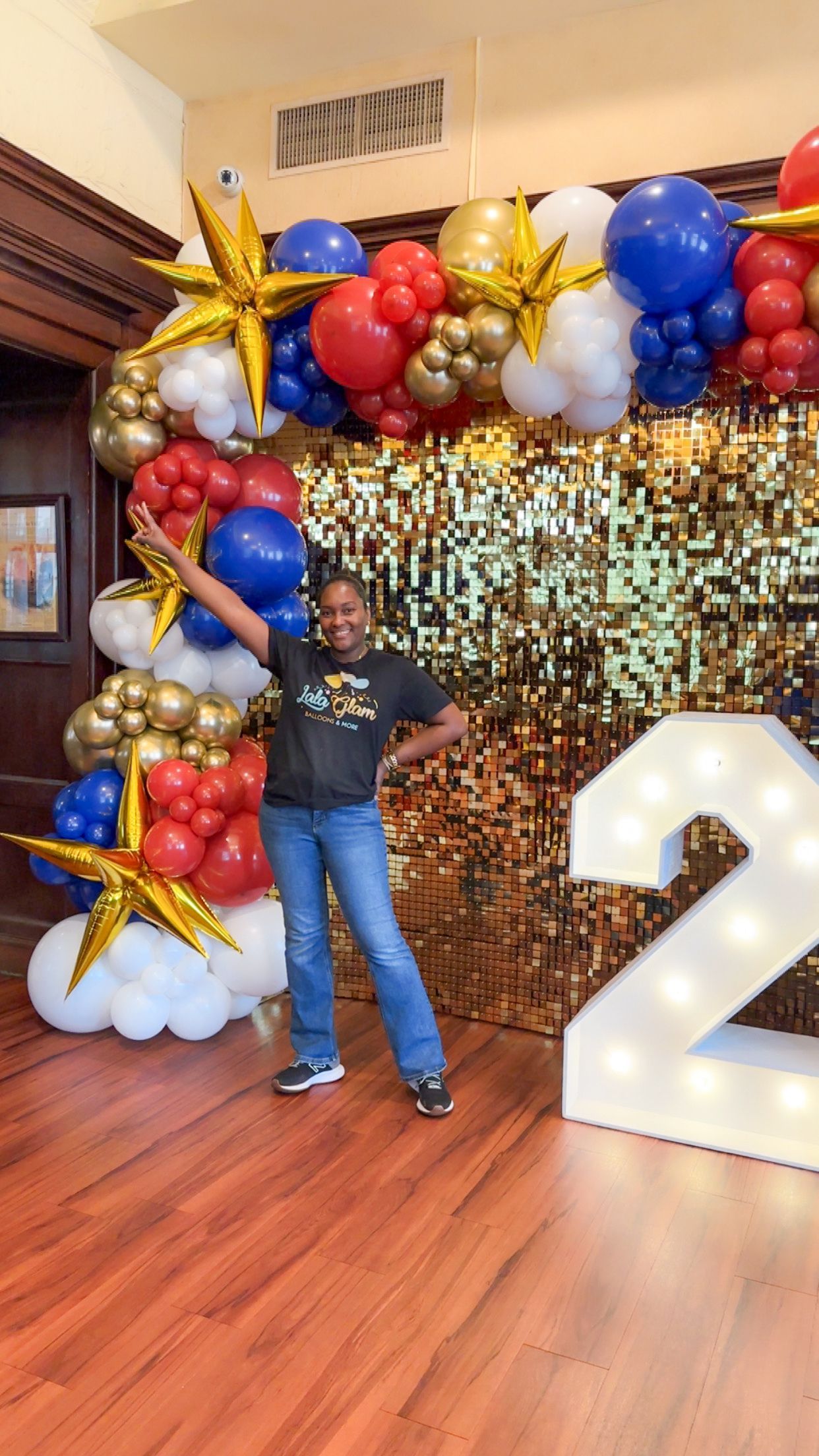 Person poses in front of gold sequin backdrop, balloon arch with starbursts and a lit-up number 2.
