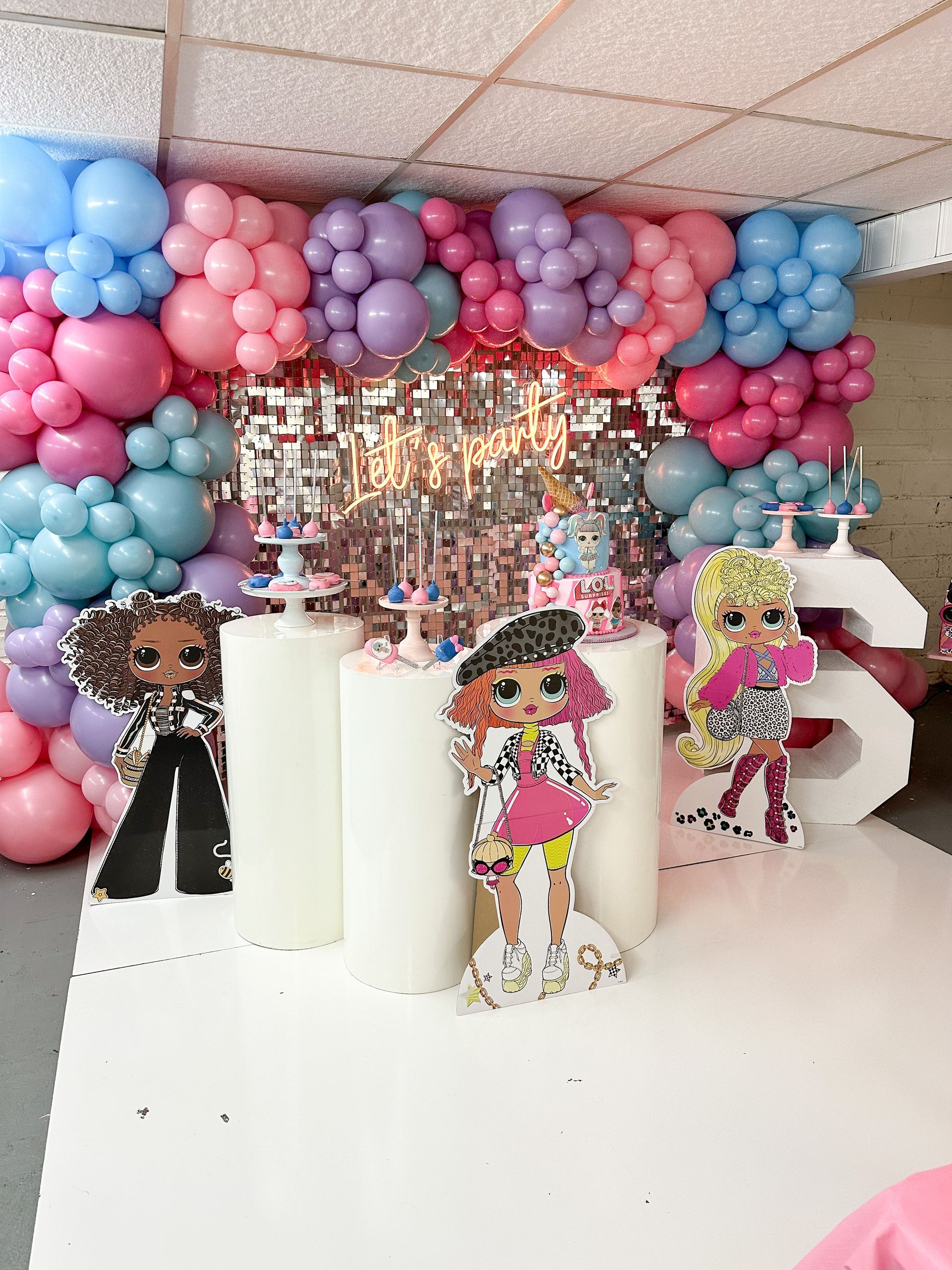 Party decorations: balloon arch in pinks/blues, dolls, white pillars, number 6, silver backdrop.