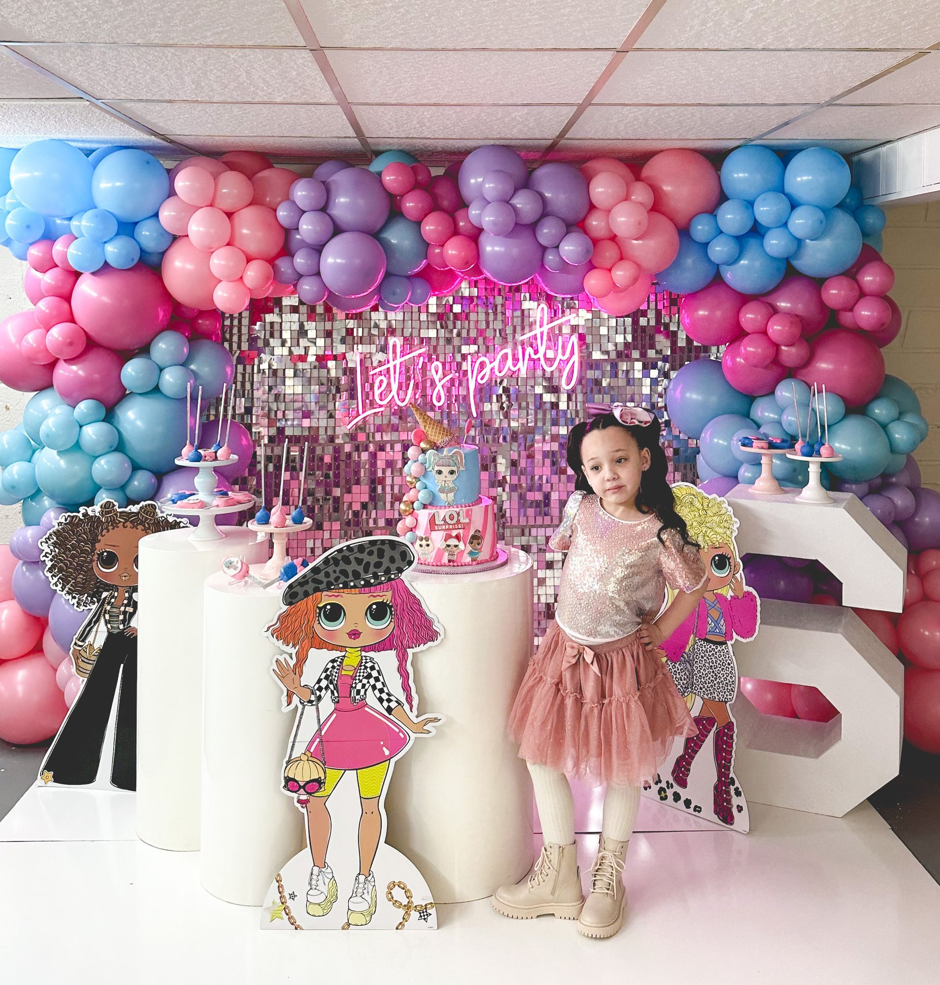 Girl in party outfit poses at a birthday celebration with balloons, cutouts, and a neon sign.