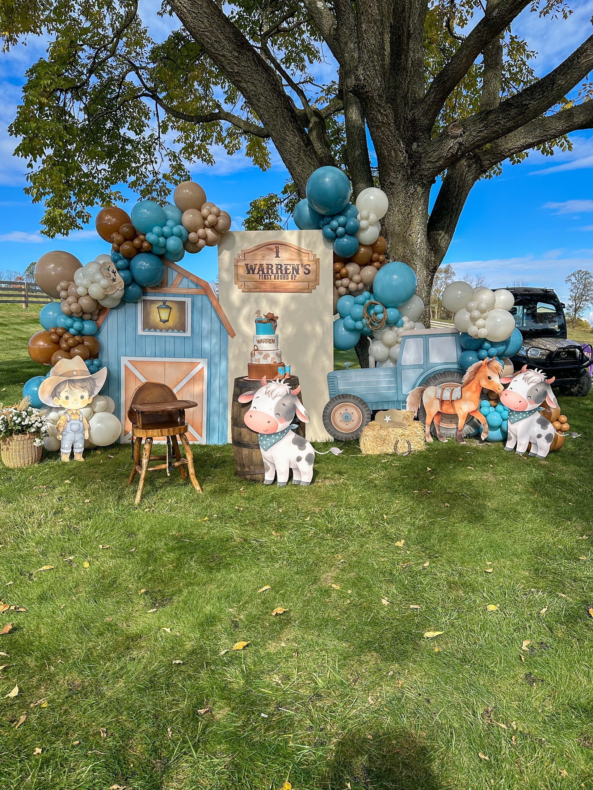 Farm-themed birthday backdrop with balloons, barn, tractor, cow and horse figurines, and a name sign on green grass.