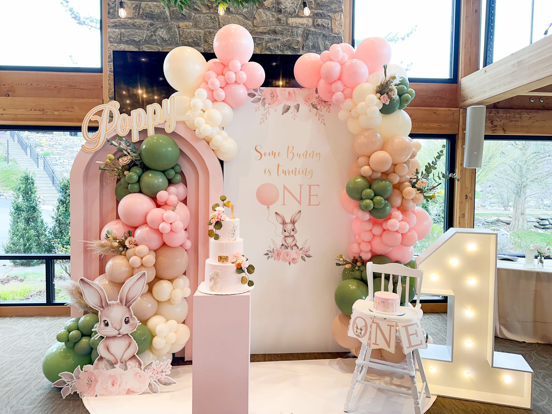 First birthday party with pink and green balloon arch, bunny decor, and a cake.