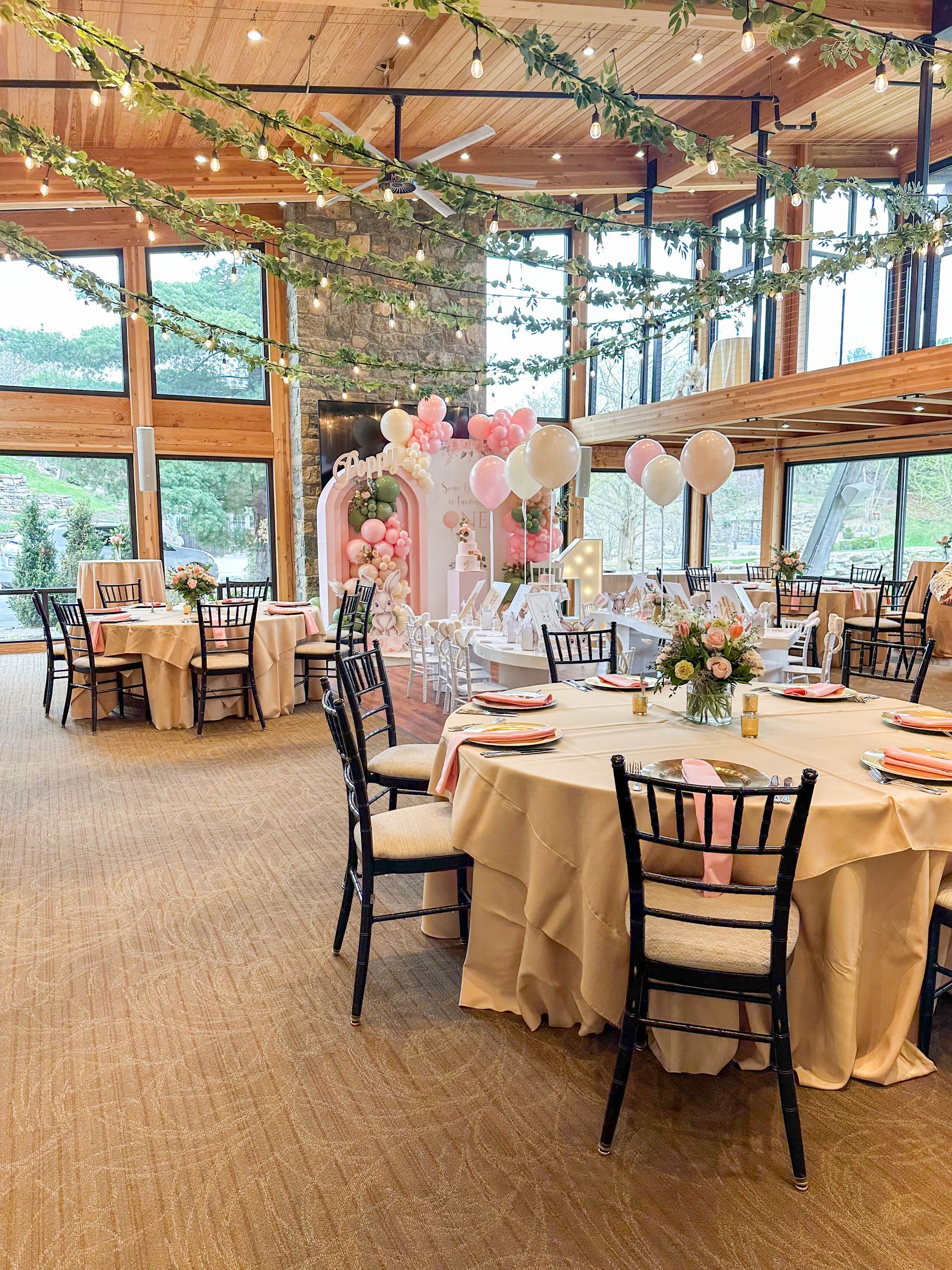 Event space decorated with pink balloons, floral arrangements, and round tables with beige linens.