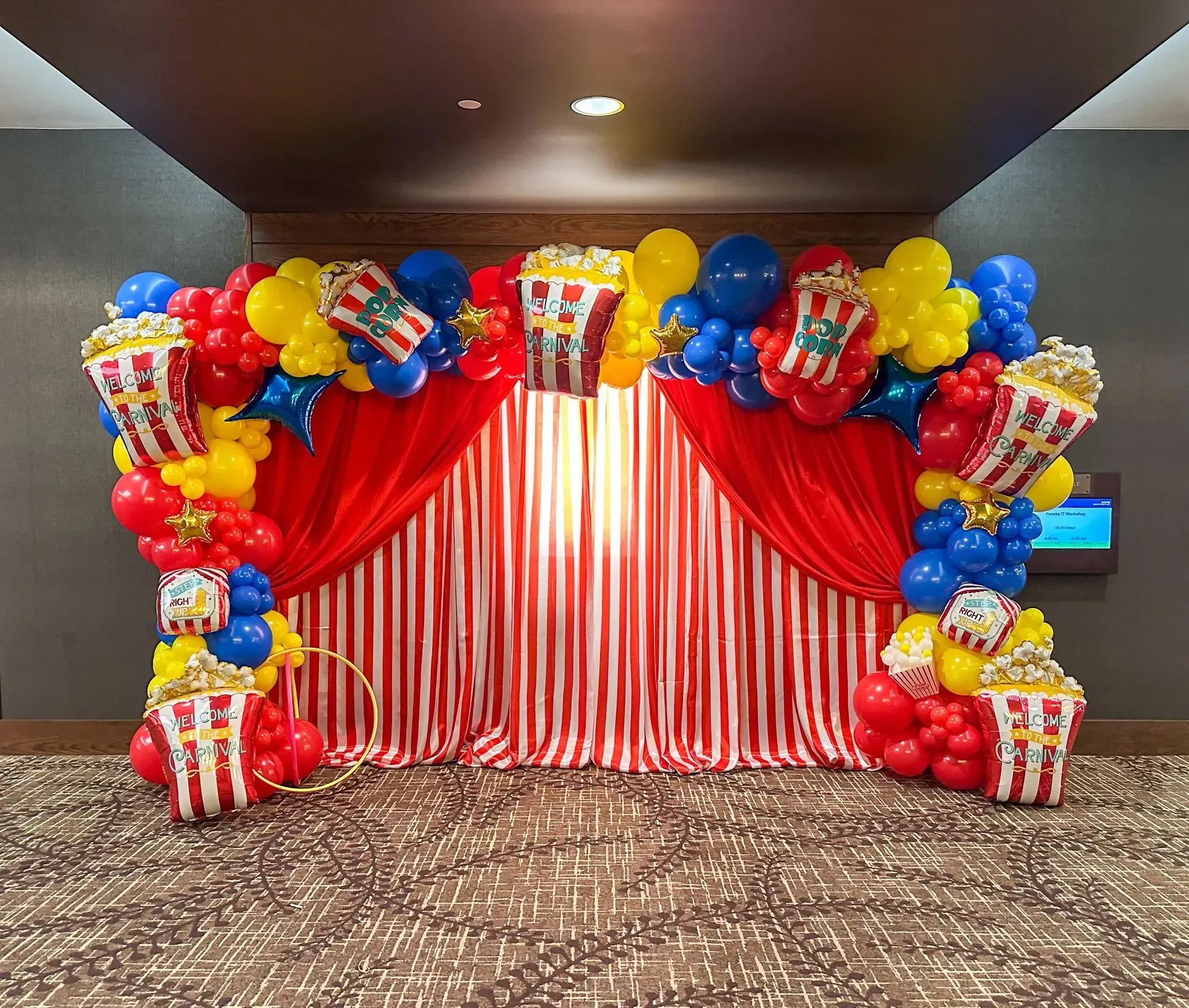 Circus-themed backdrop with red/white striped curtains, popcorn balloons, and colorful balloon arches.