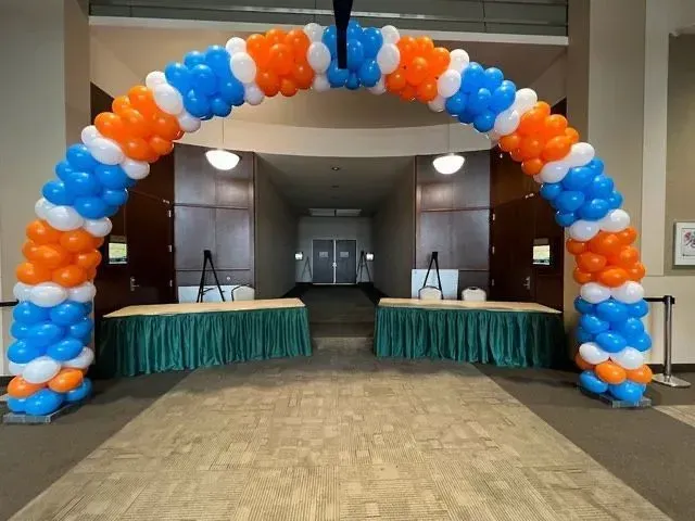 Balloon arch in blue, white, and orange over two draped tables in a hallway.