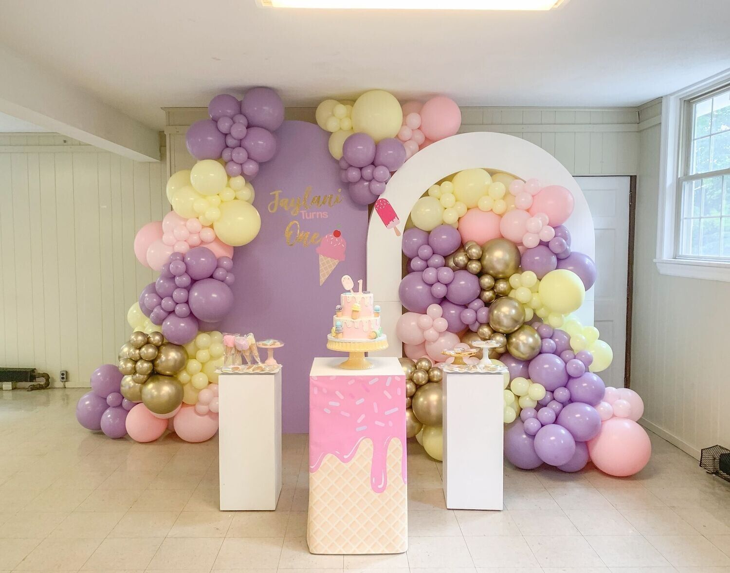 Birthday party backdrop with balloons in lavender, yellow, pink, and gold, featuring a cake and podiums.