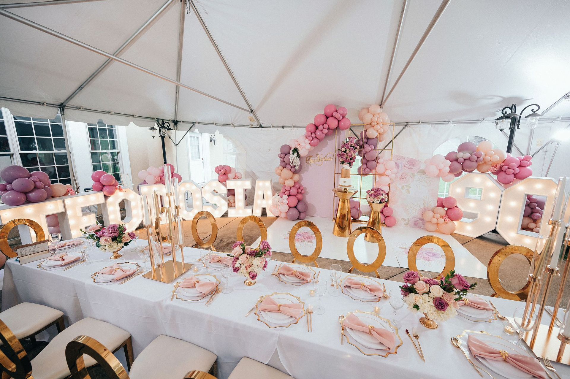 Party table decorated in pink and gold, under a tent. Balloons, florals, and a light-up