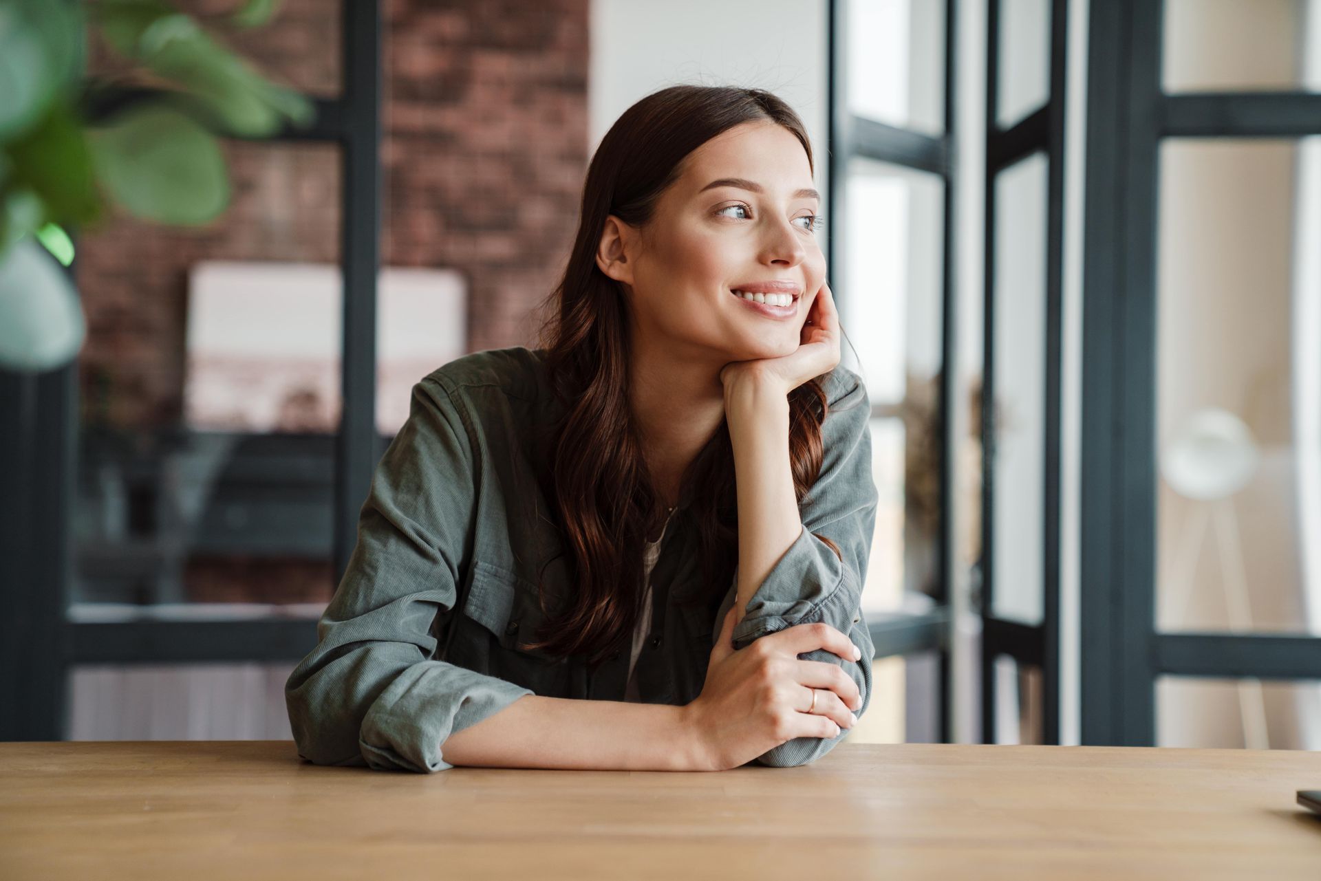 A woman is sitting at a table with her hand on her chin and looking out the window.