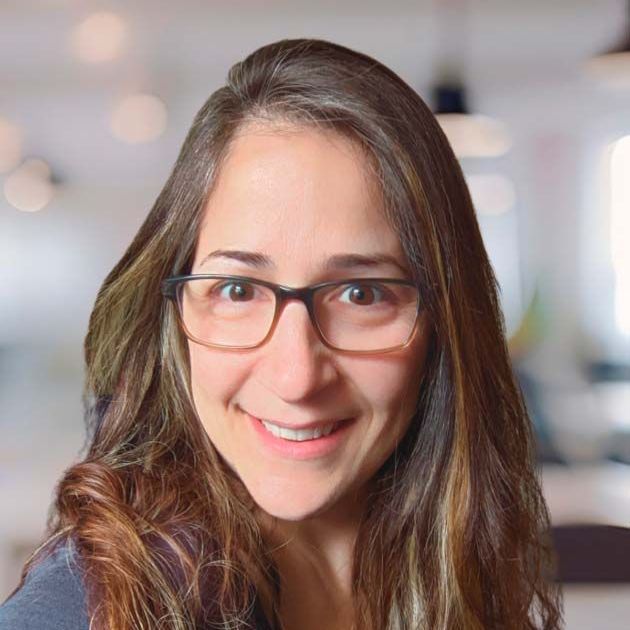 A woman is sitting on a couch talking to another woman while holding a laptop.