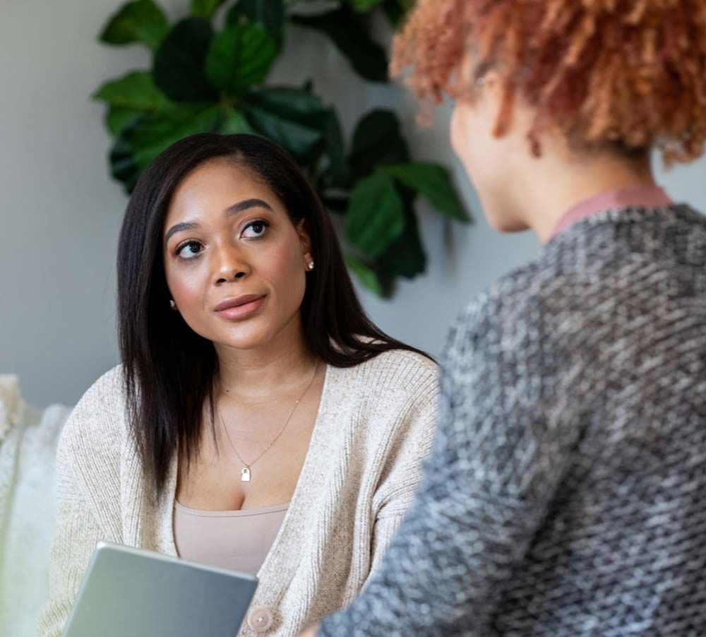 A woman is sitting on a couch talking to another woman while holding a laptop.