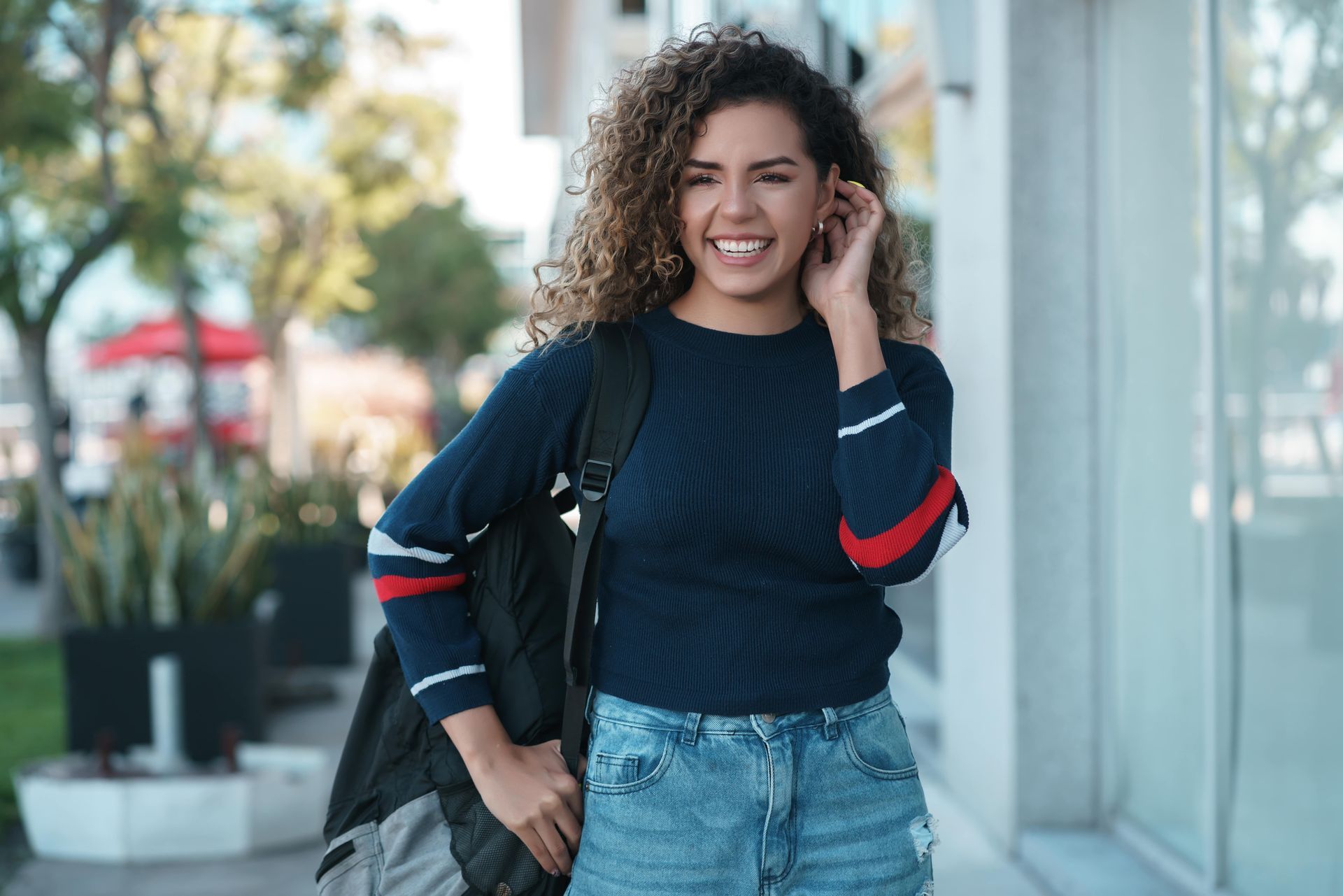 A woman with curly hair is standing on a sidewalk with a backpack.