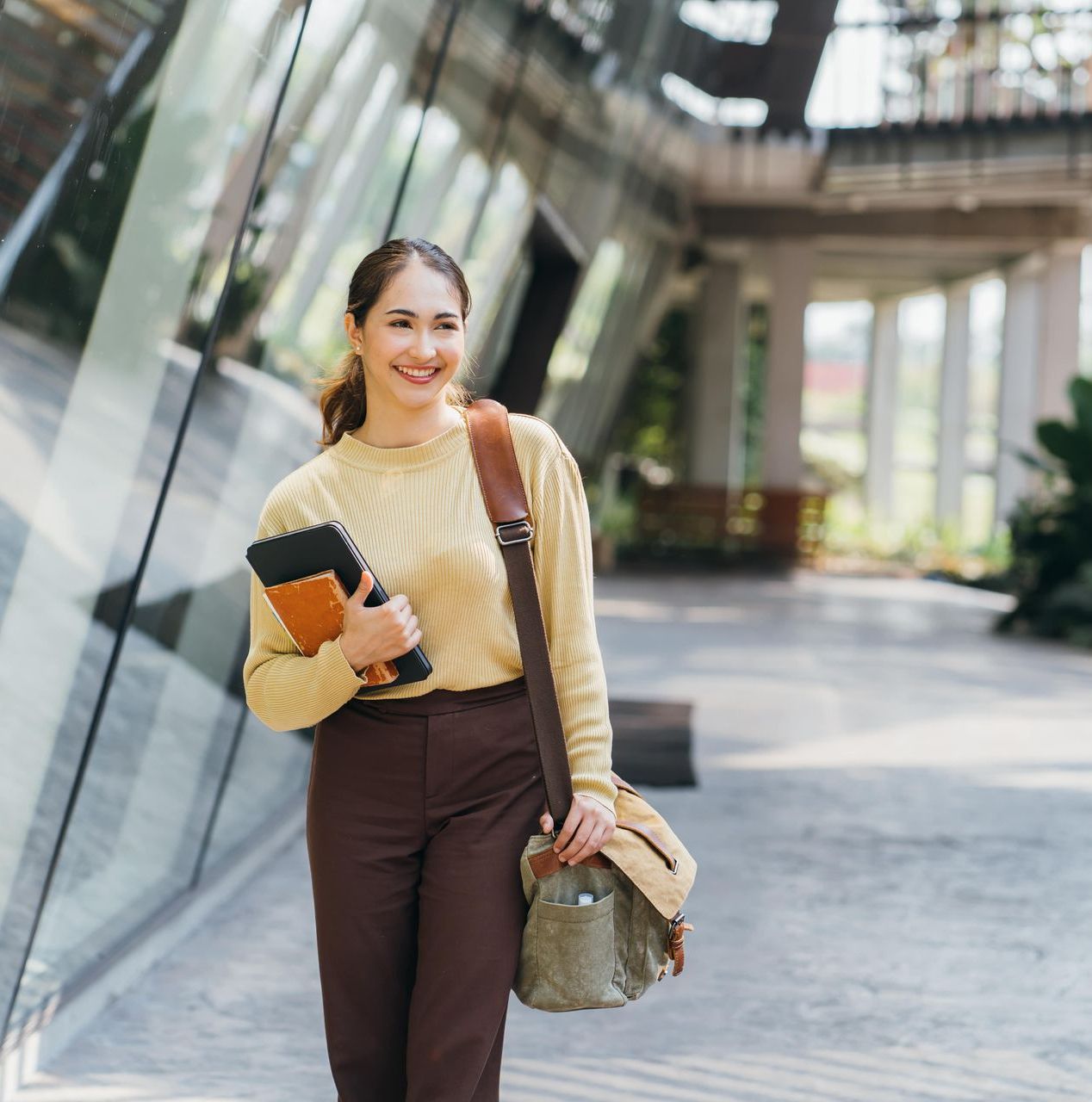 A woman is walking down a hallway holding a book and a bag