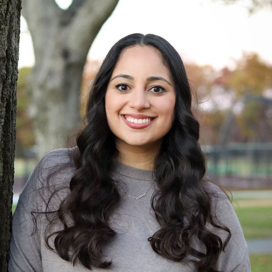 A woman is sitting on a couch talking to another woman while holding a laptop.