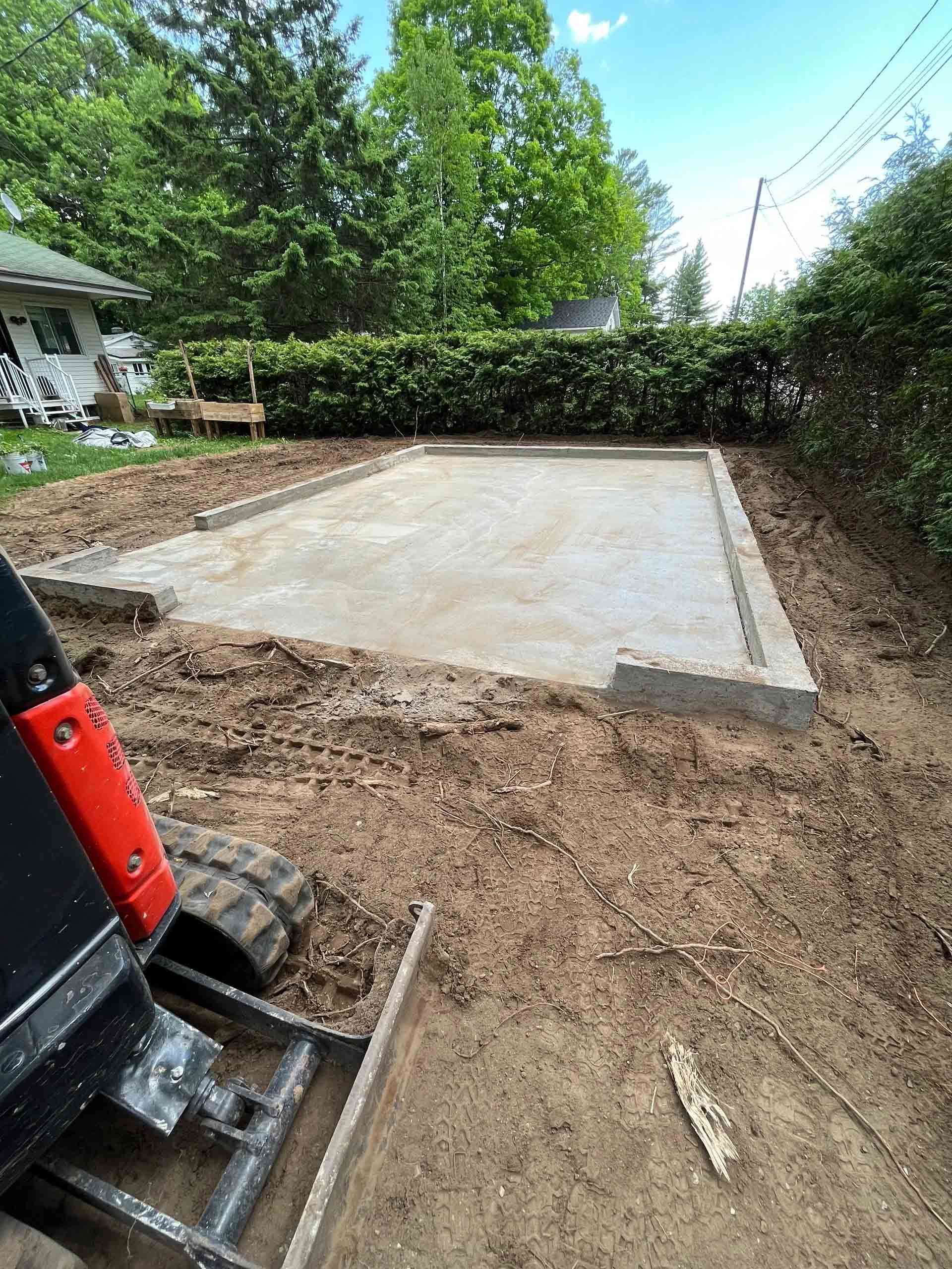 Dalle de béton en construction dans une cour arrière, entourée de terre et d'arbres à proximité.