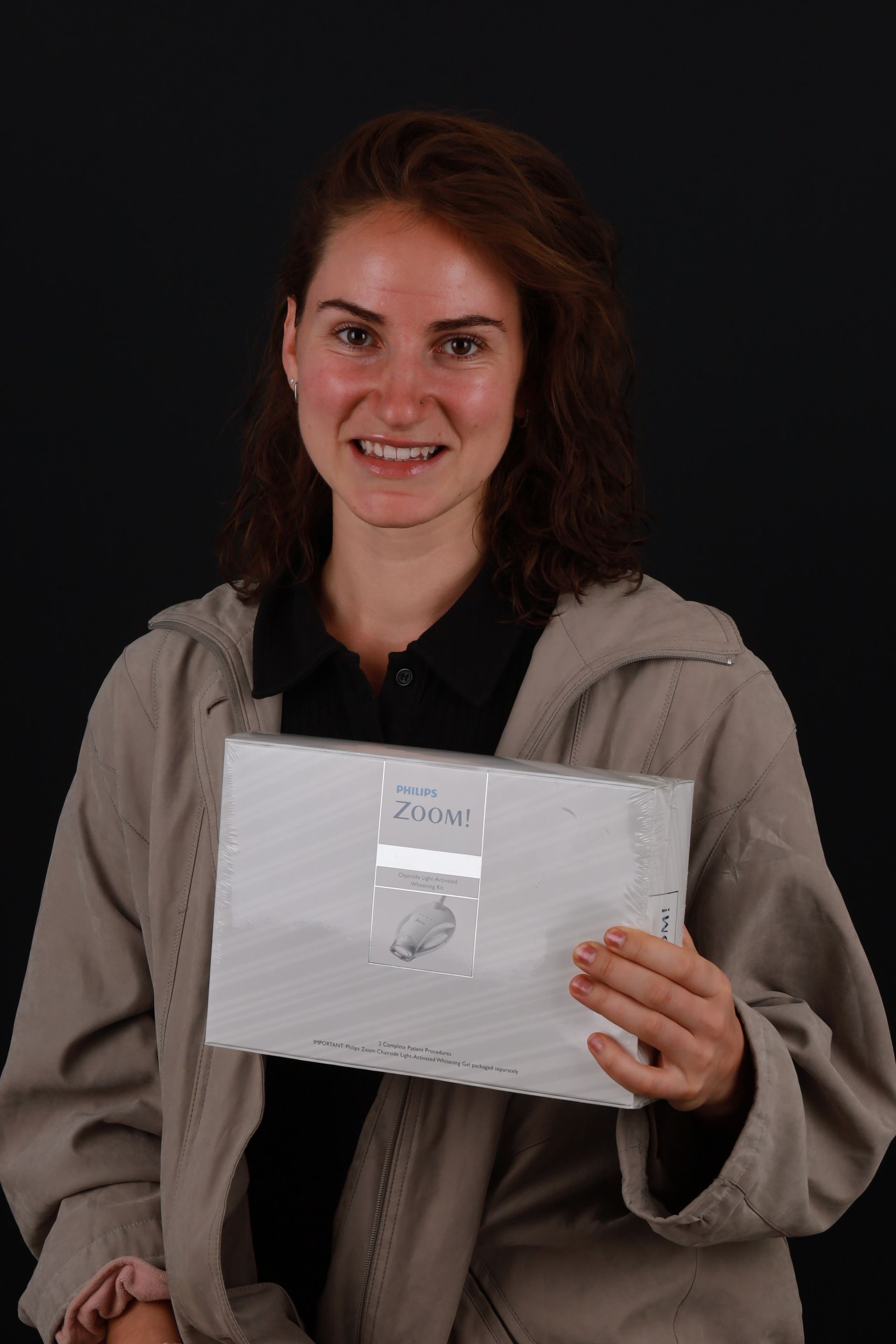 Woman in tan jacket smiles, holding sealed envelope with visible writing against a black background.