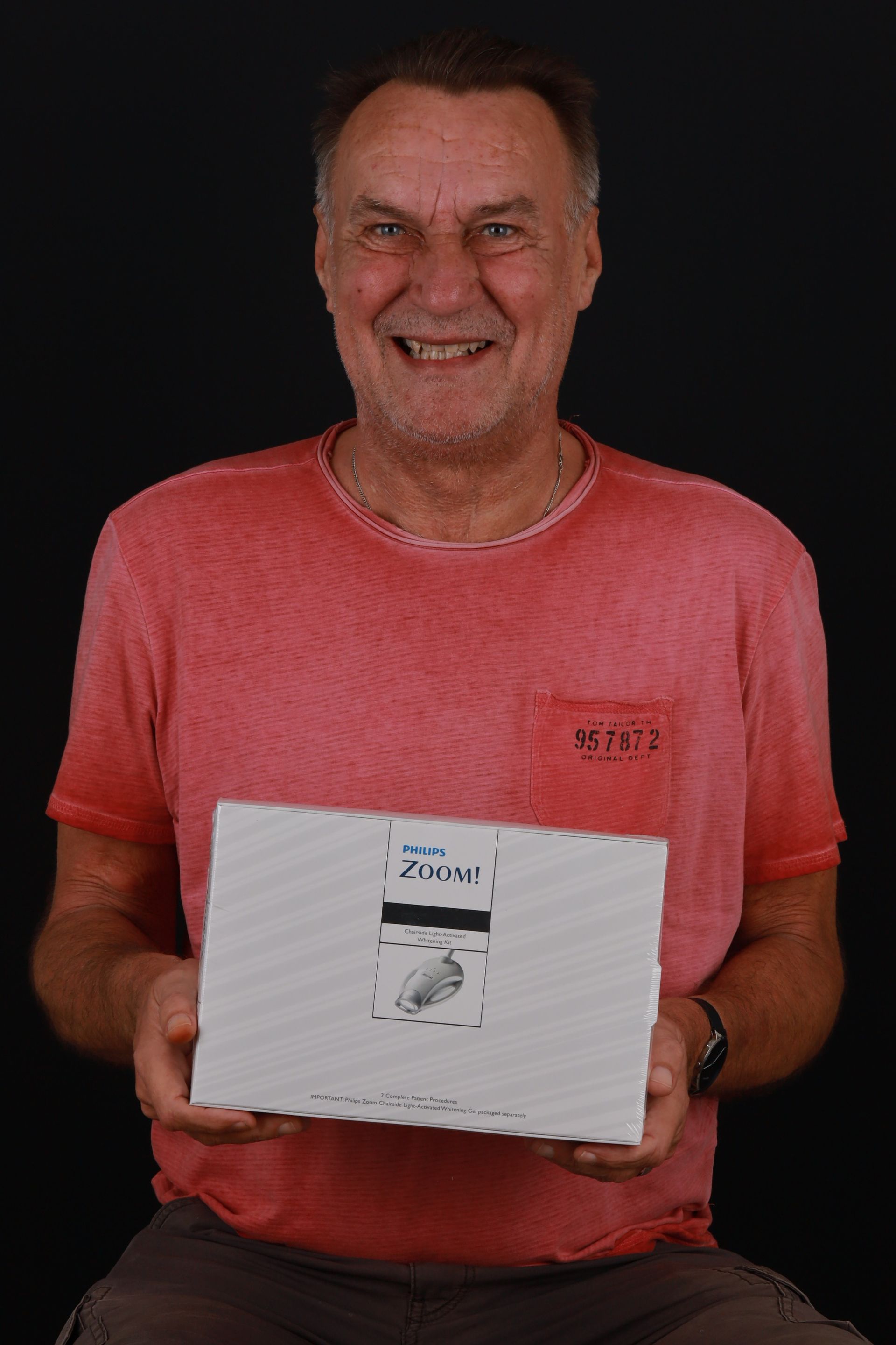 Smiling older man in red shirt holding a white box with a drawing, against a dark background.