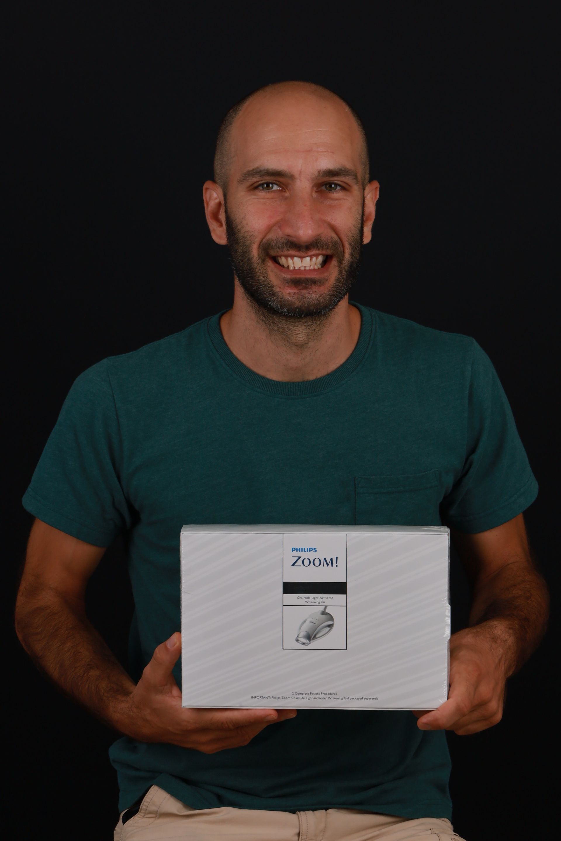 Man with a beard smiles, holding a white box against a black background.