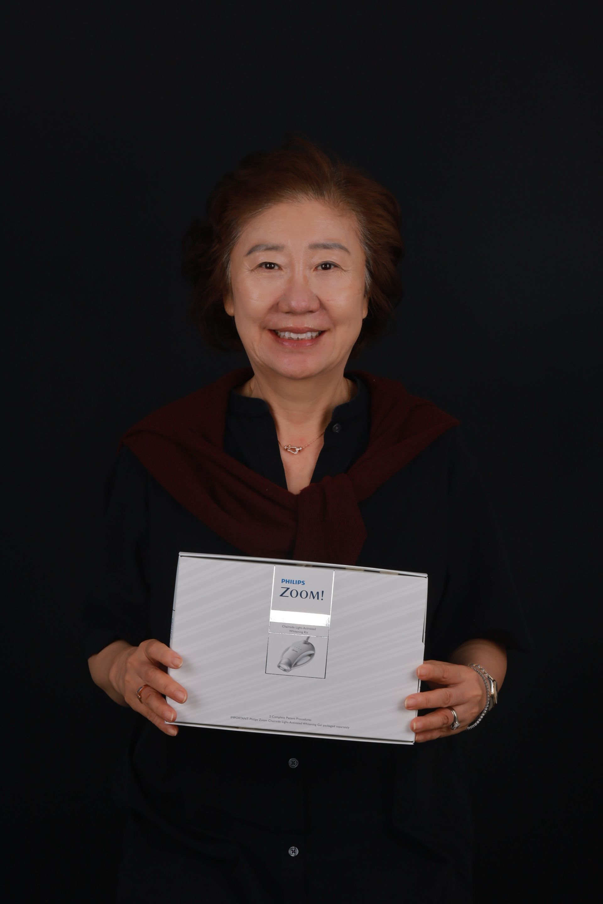 Woman with dark hair, smiling, holding a white box against a black background.