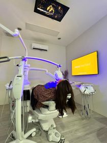 Dental chair with a mannequin patient wearing a dark wig, in a brightly lit dentist's office.
