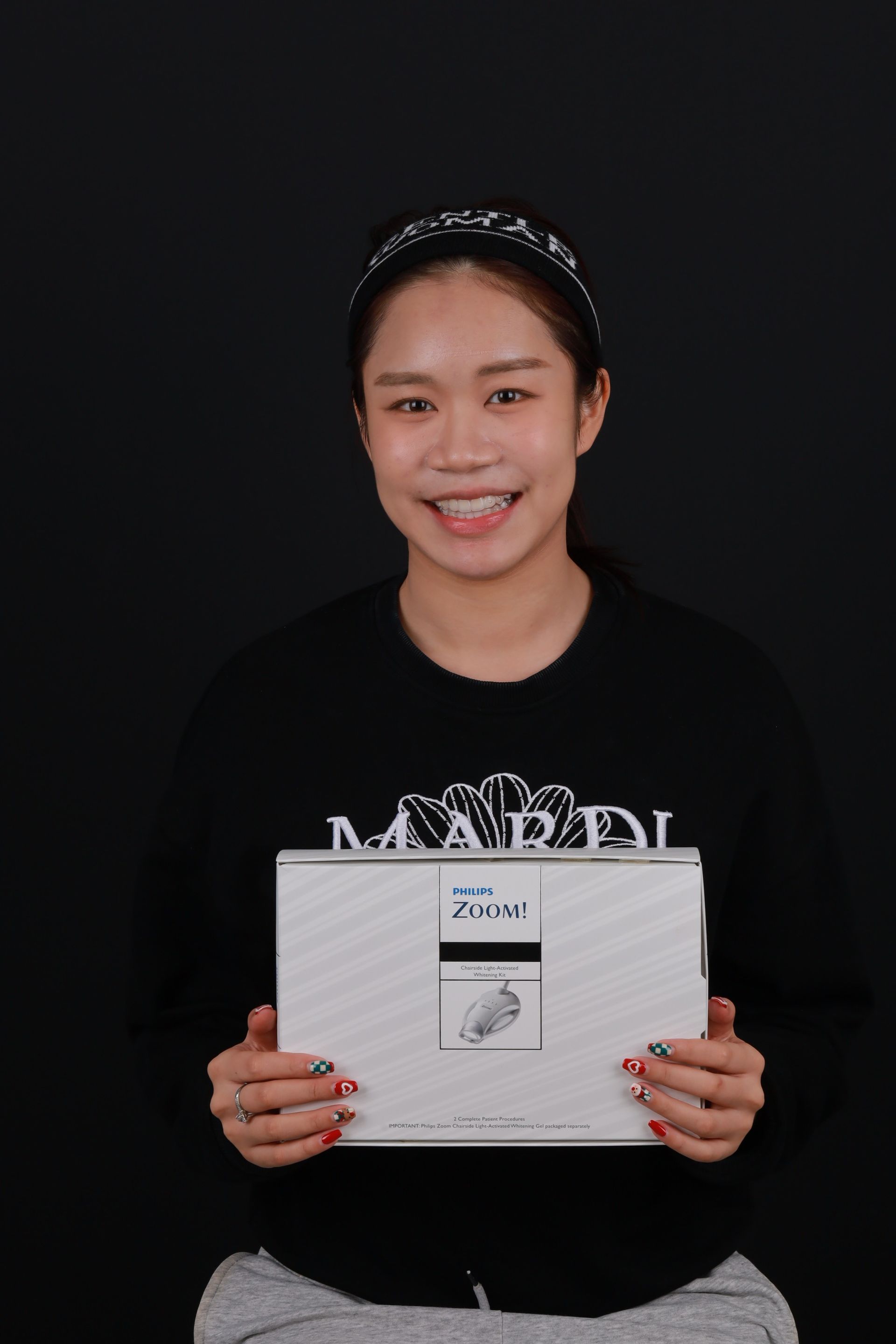 Woman with braces smiles, holding a white box against a black background.