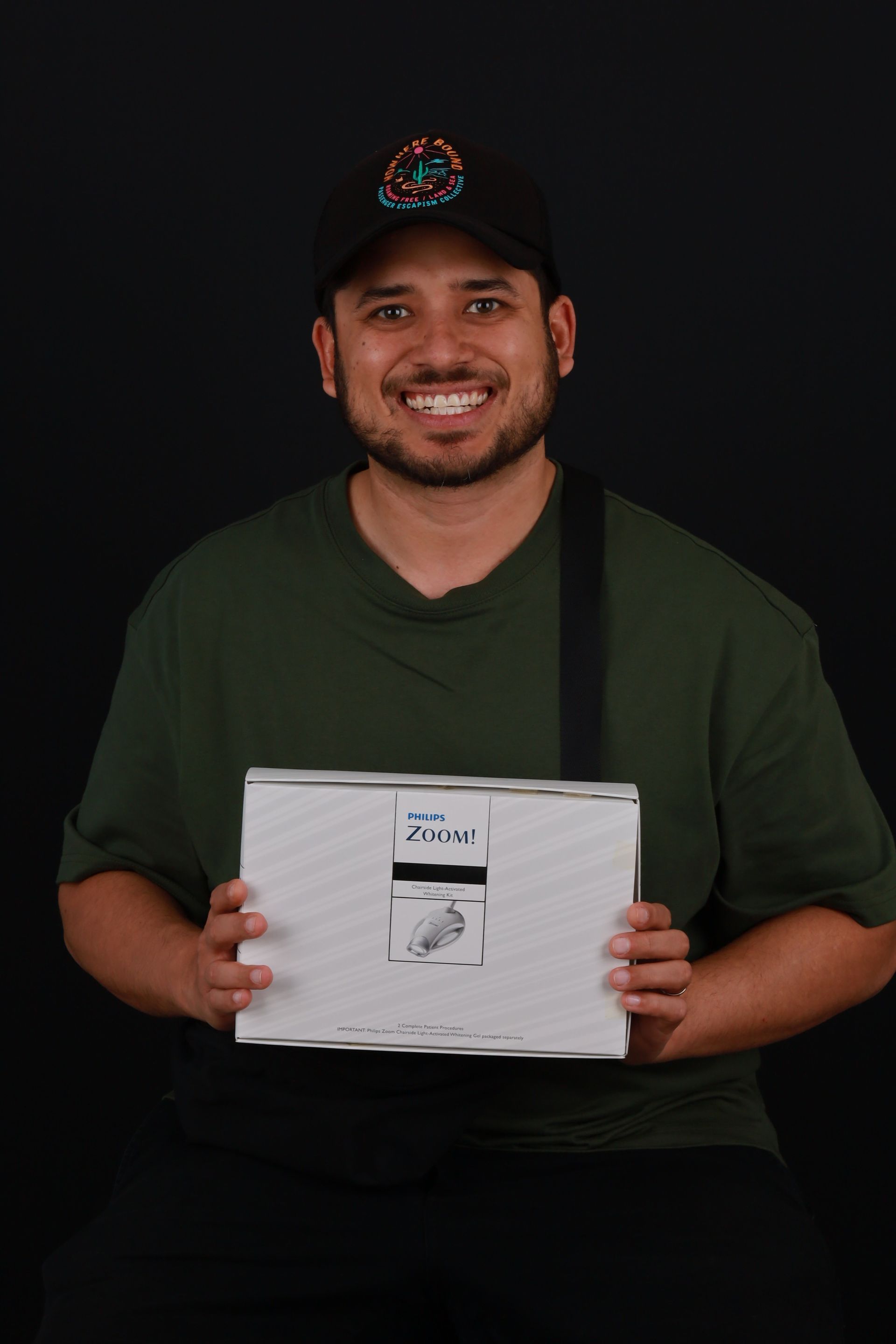 Man holding a white box, smiling, wearing a cap and green shirt against a black background.
