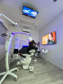 Dental clinic interior: patient in chair, undergoing teeth whitening treatment under blue light, TV displays.