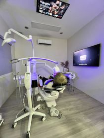 Dental office interior: person in chair, undergoing procedure, with bright equipment; screens on wall and ceiling.