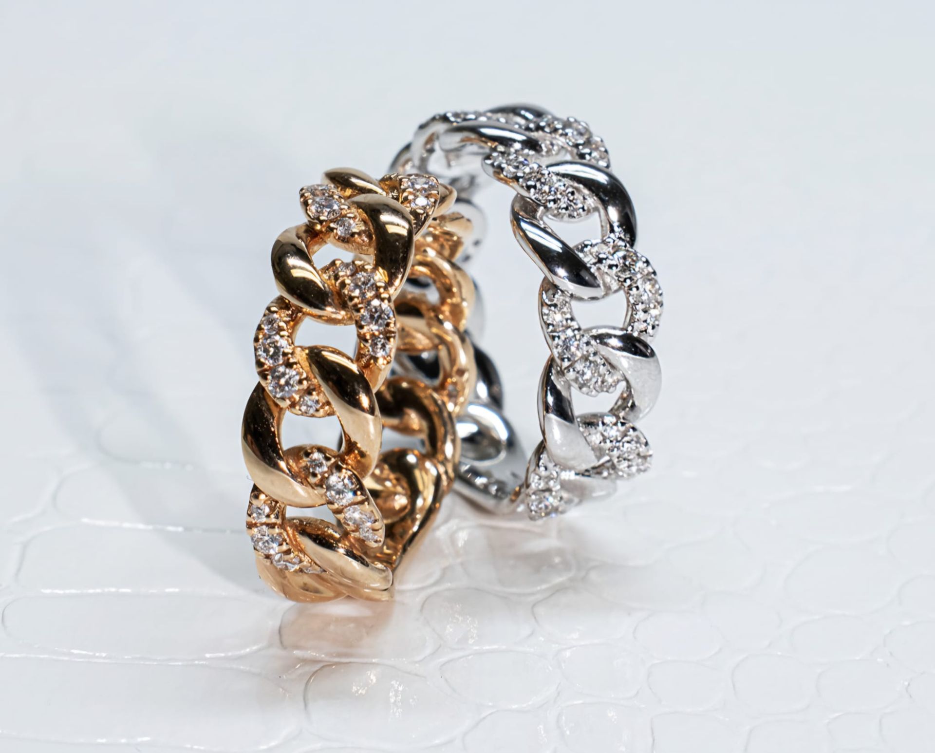 Due anelli tempestati di diamanti, uno in oro e uno in argento, sono appoggiati su una superficie bianca testurizzata.