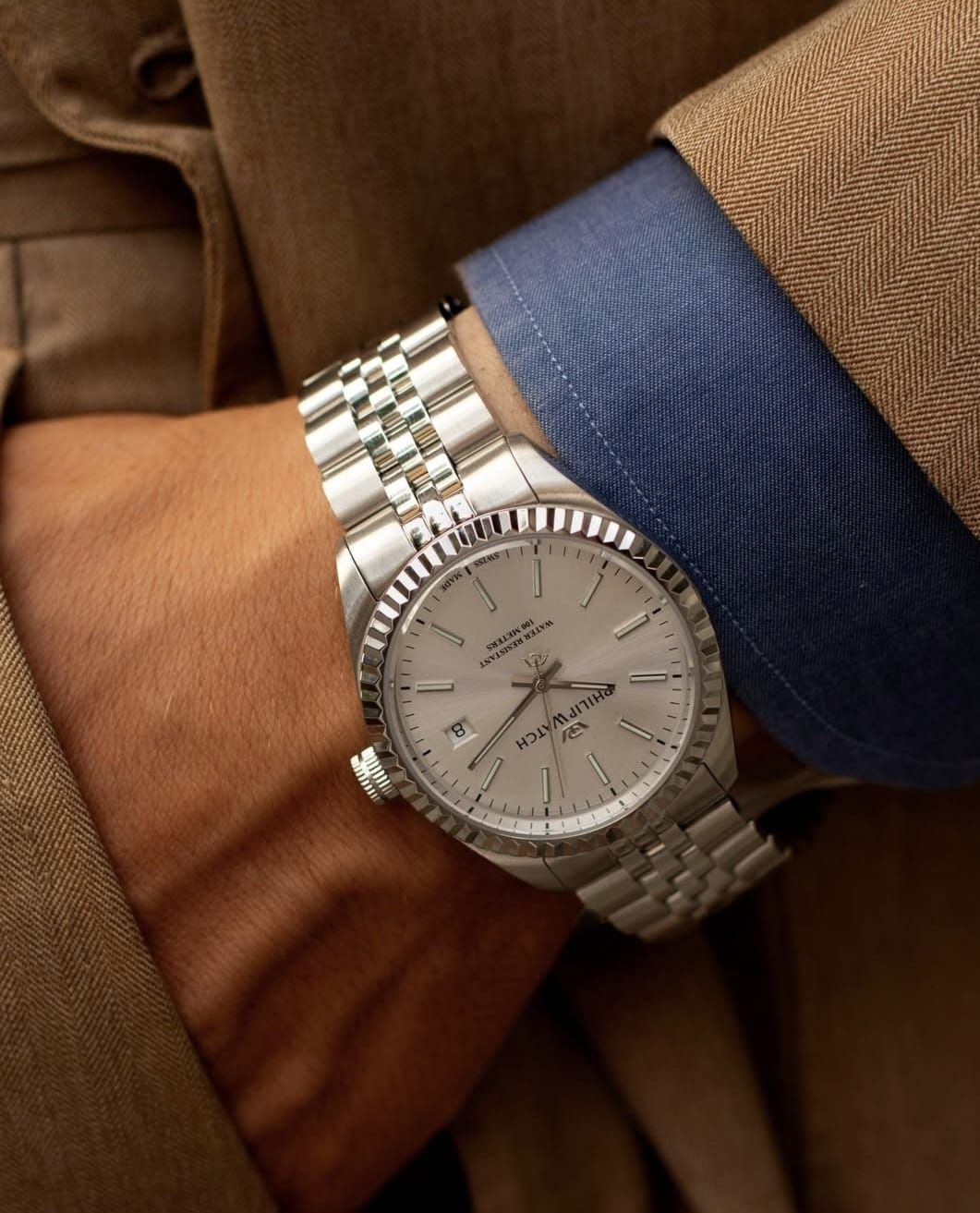 Orologio d'argento al polso; al braccio una camicia blu con un blazer marrone chiaro.