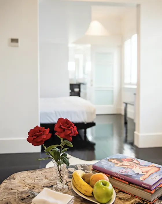 Table with fruit, roses, and a book in a modern room with a view of a bed.