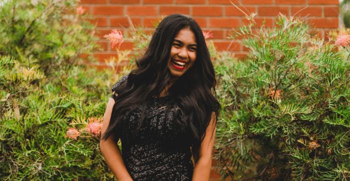 Woman laughing in front of leafy green bush and brick wall; she's wearing a black sequin top.