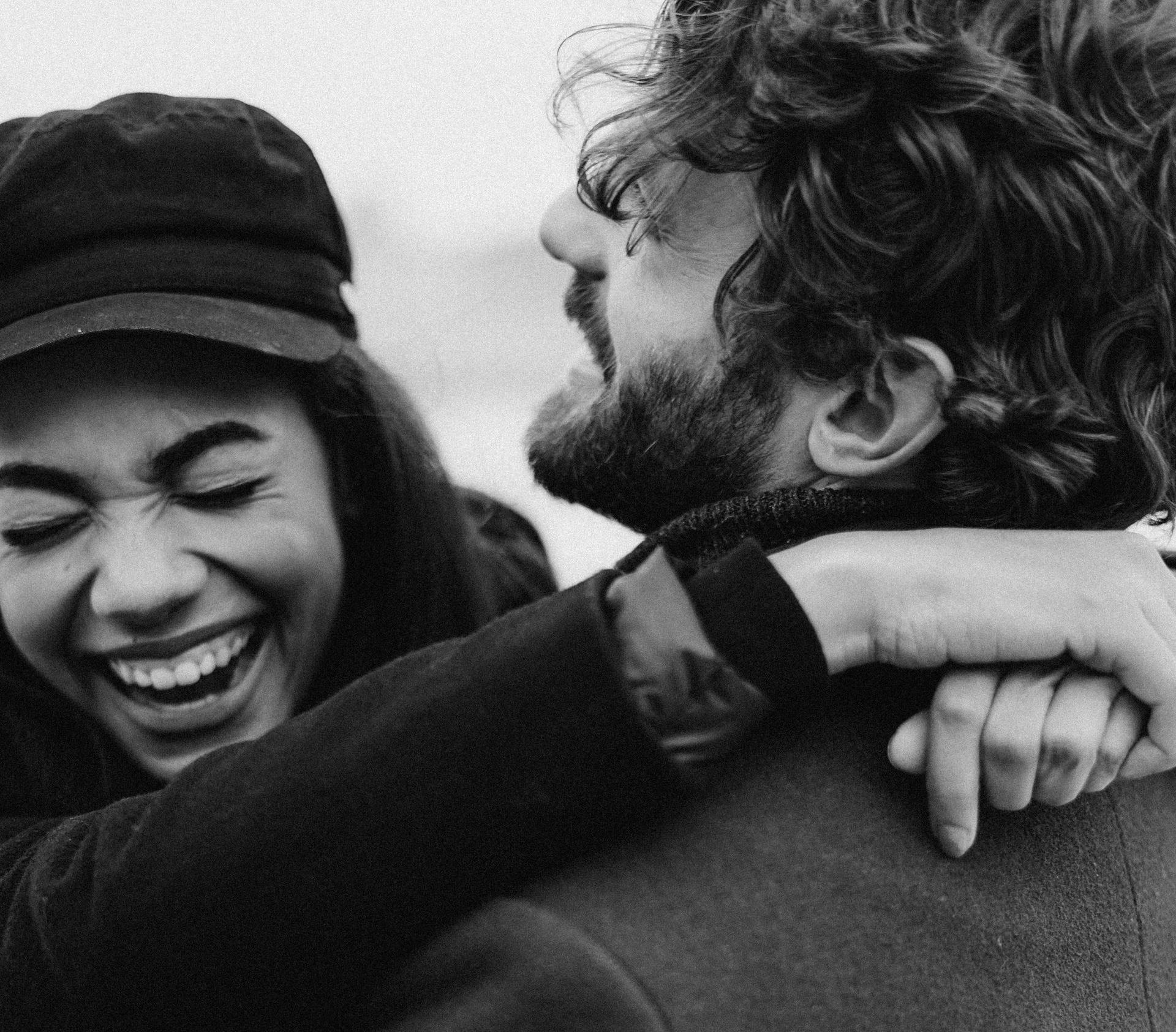 Woman in hat laughing and hugging person with beard.