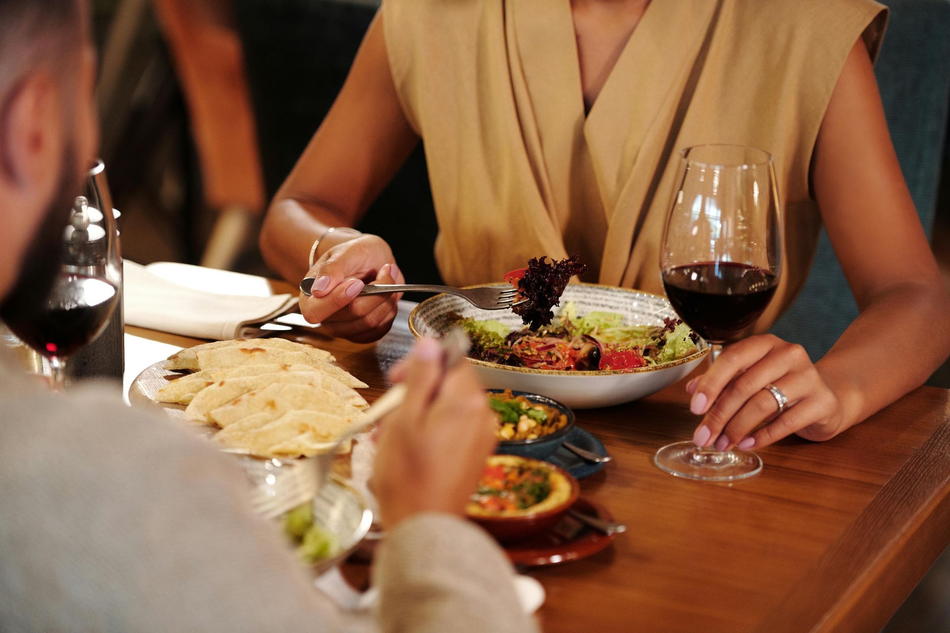 Couple dining in a restaurant; person holding a wine glass and fork. Salad and other dishes on the table.