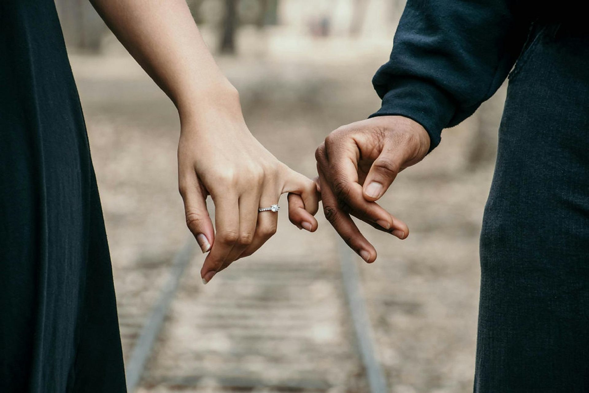 Couple holding pinky fingers, one wearing an engagement ring, against blurred outdoor background.