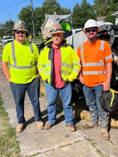 Three construction workers are posing for a picture on a sidewalk.