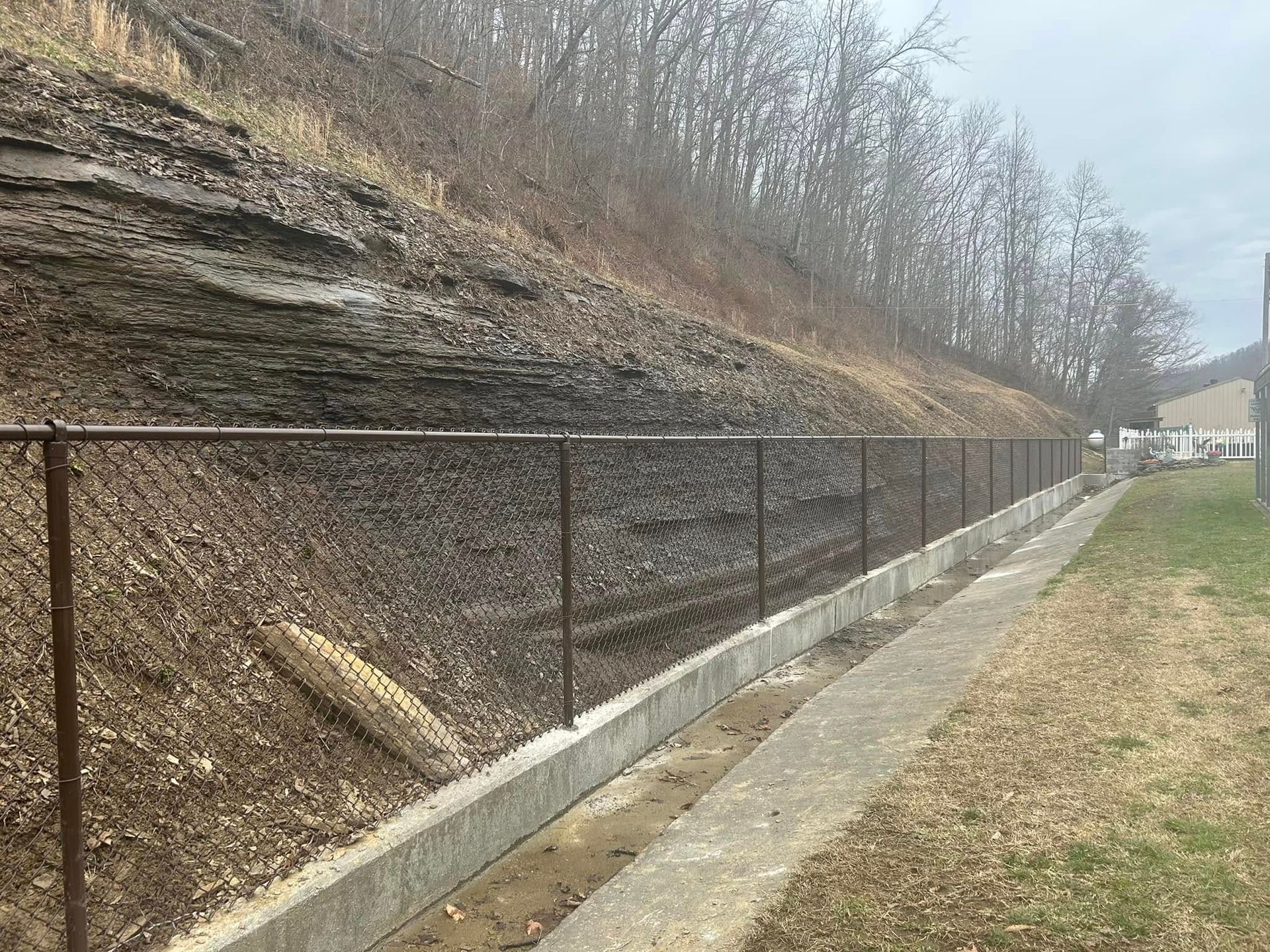 A fence along a sidewalk next to a hill with trees in the background.
