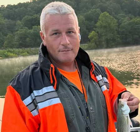 A man in an orange jacket is holding a fish in front of a lake.