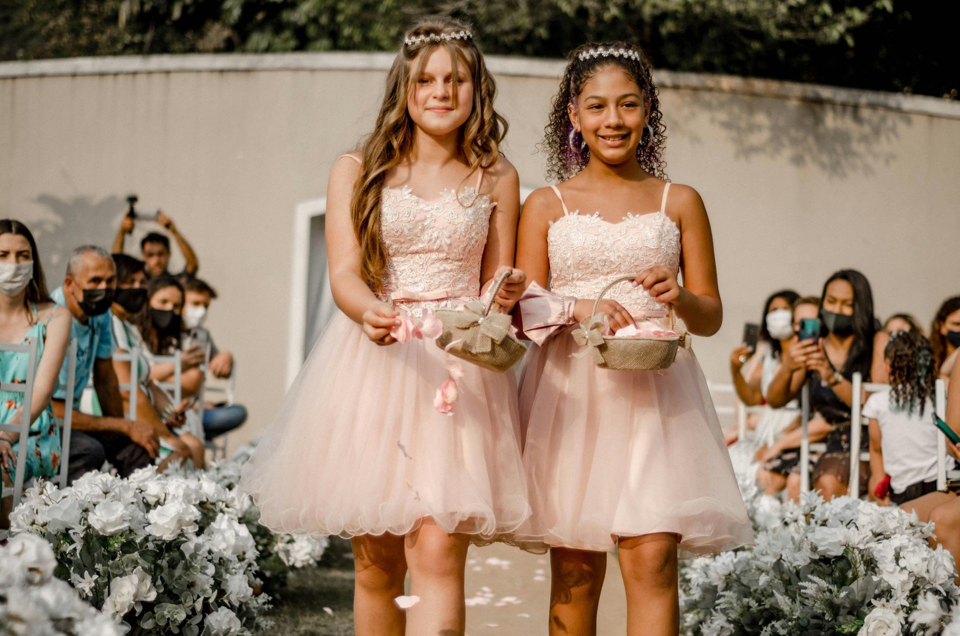 Two girls in pink dresses walking, scattering flower petals. Outdoor wedding setting.