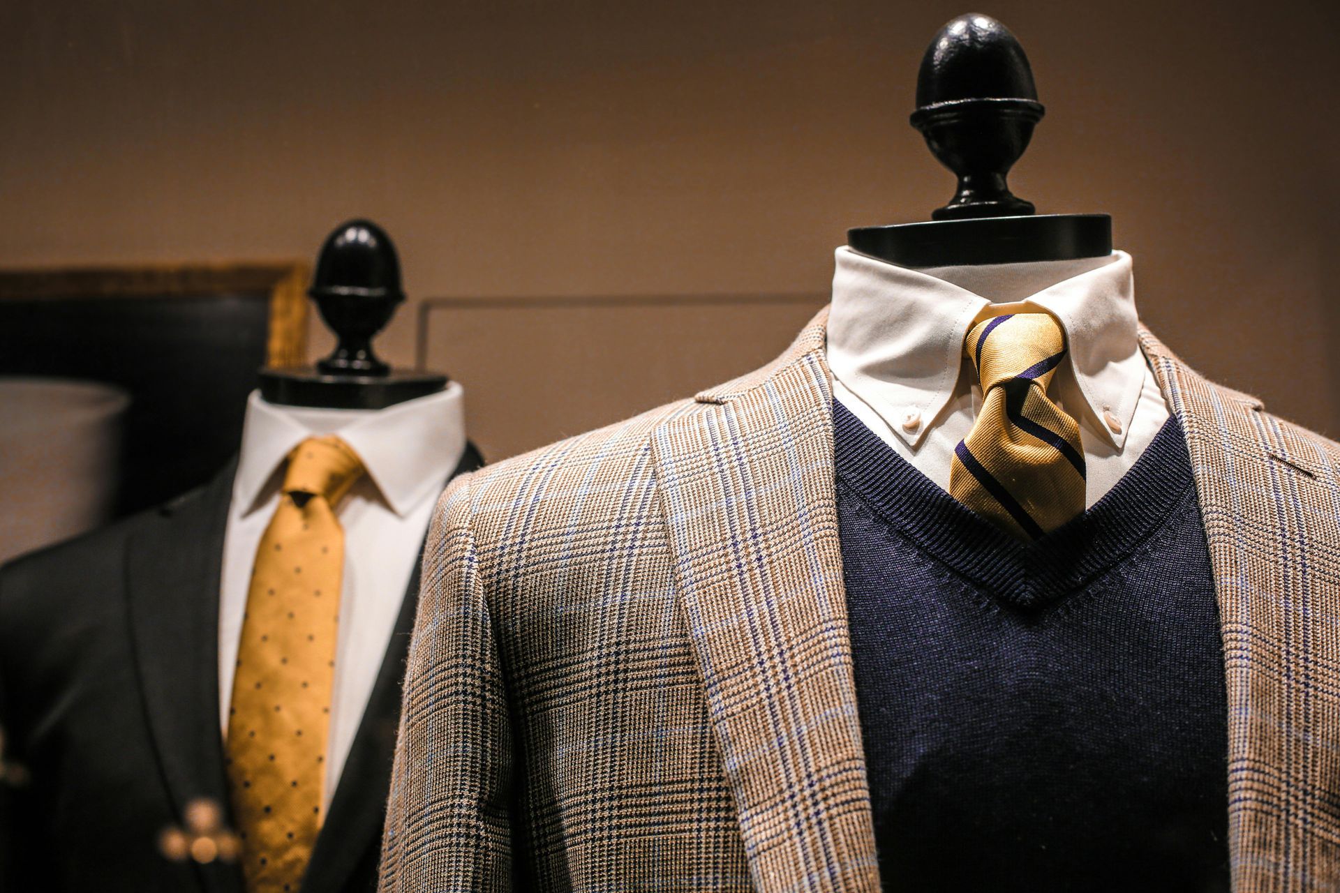Two mannequins displaying suits, one black with gold tie, the other with a patterned jacket and navy sweater.